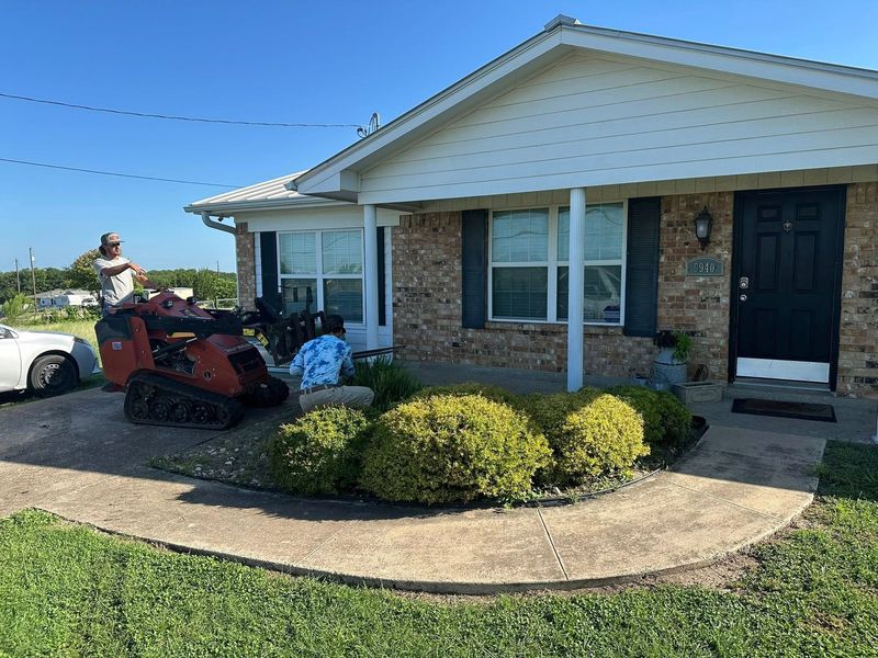 Single-story brick house with a curved walkway, red lawn mower, and trimmed bushes under a clear blue sky