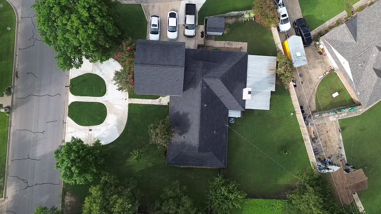 Aerial view of a suburban house with driveway, lawn, and parked cars beside it.