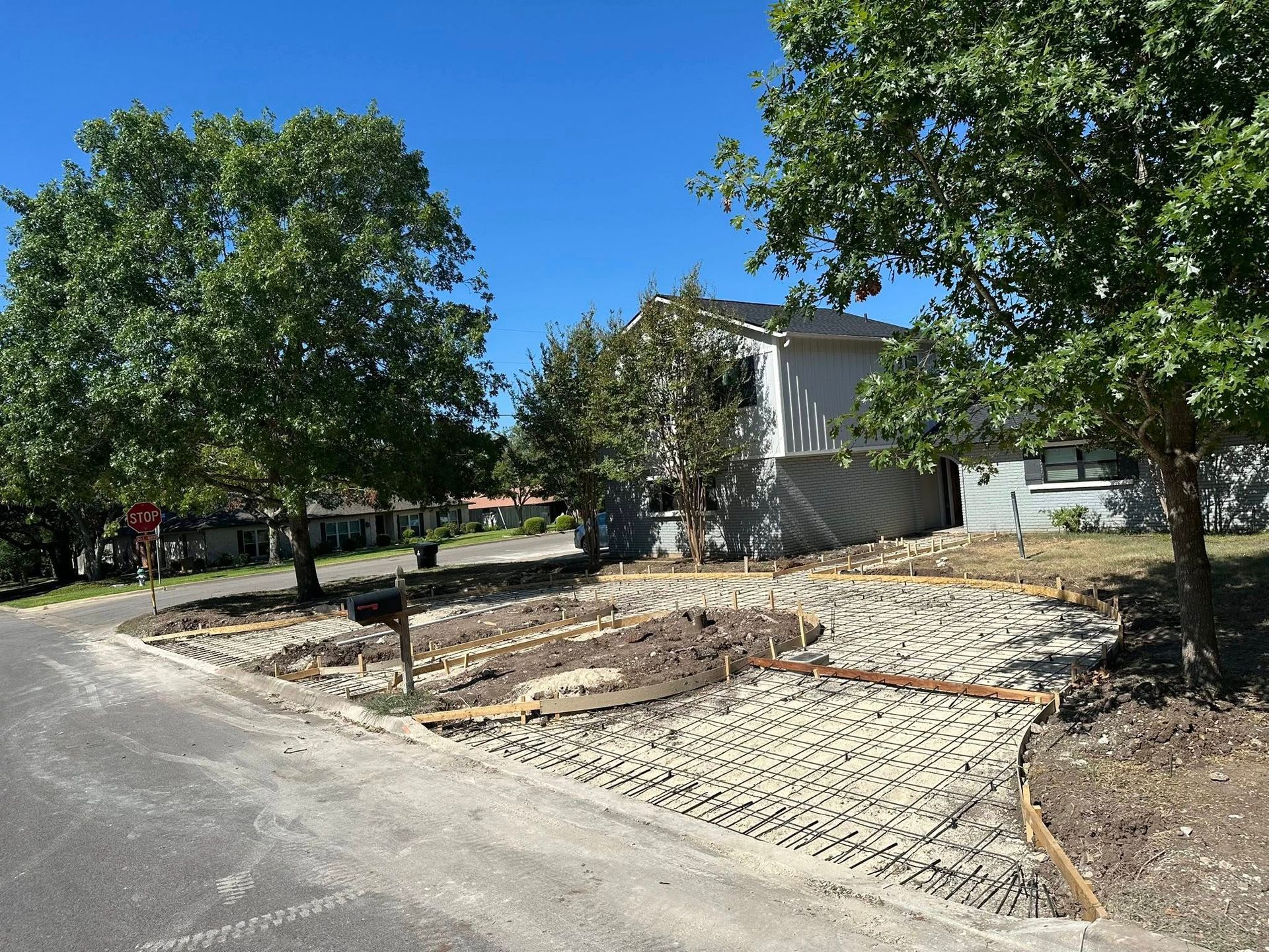 Quiet suburban street with a sandy front yard, trees, and a house under a clear blue sky