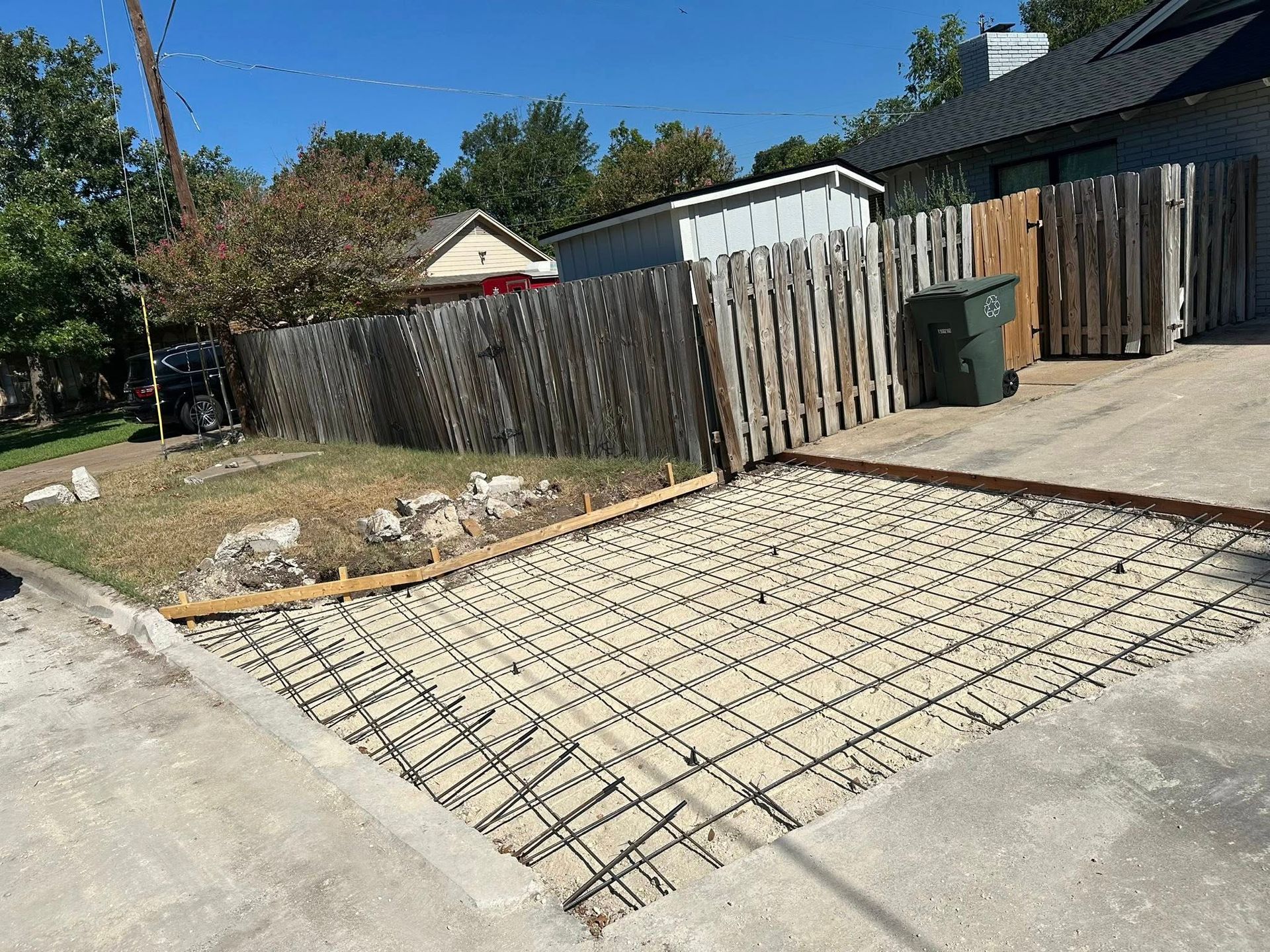 Rebar grid laid across a driveway entrance beside a wooden fence and sidewalk under a clear blue sky