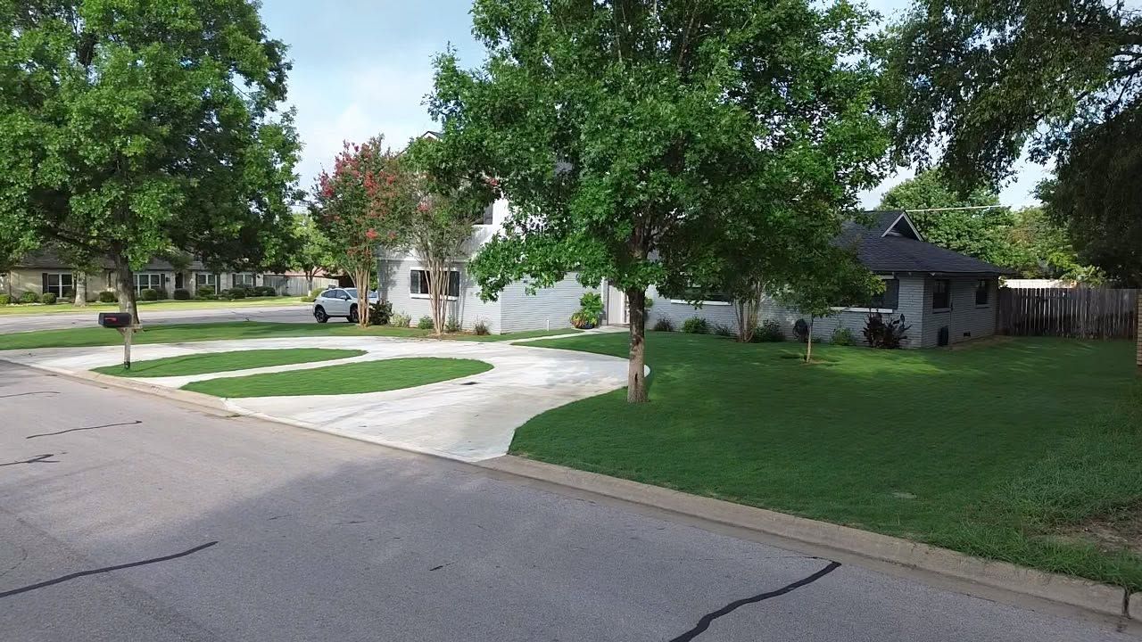 Quiet suburban street with a white house, green lawn, trees, and a driveway under bright daylight