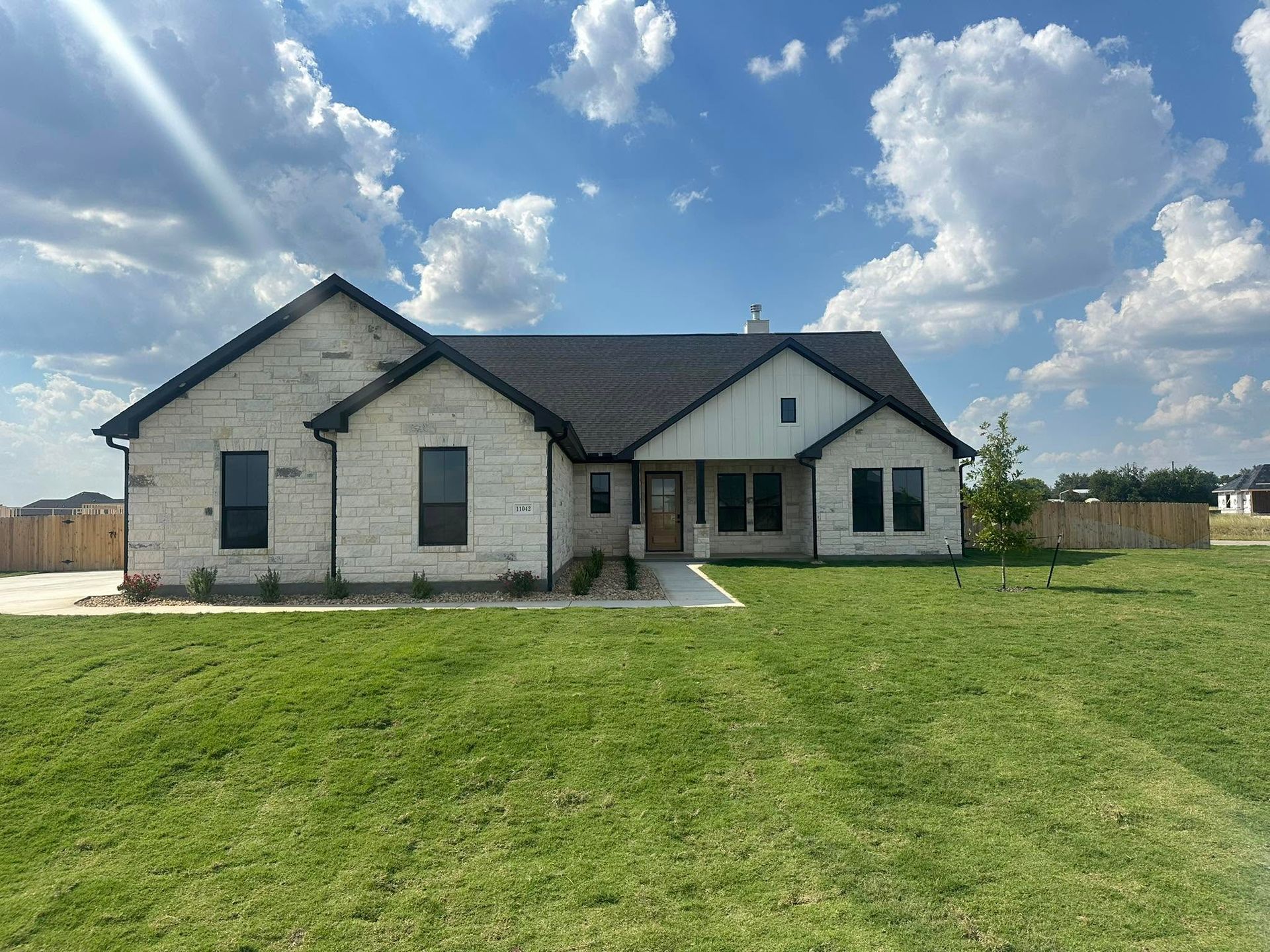 Single-story stone house with dark roof and front porch on a large grassy lot under a partly cloudy sky