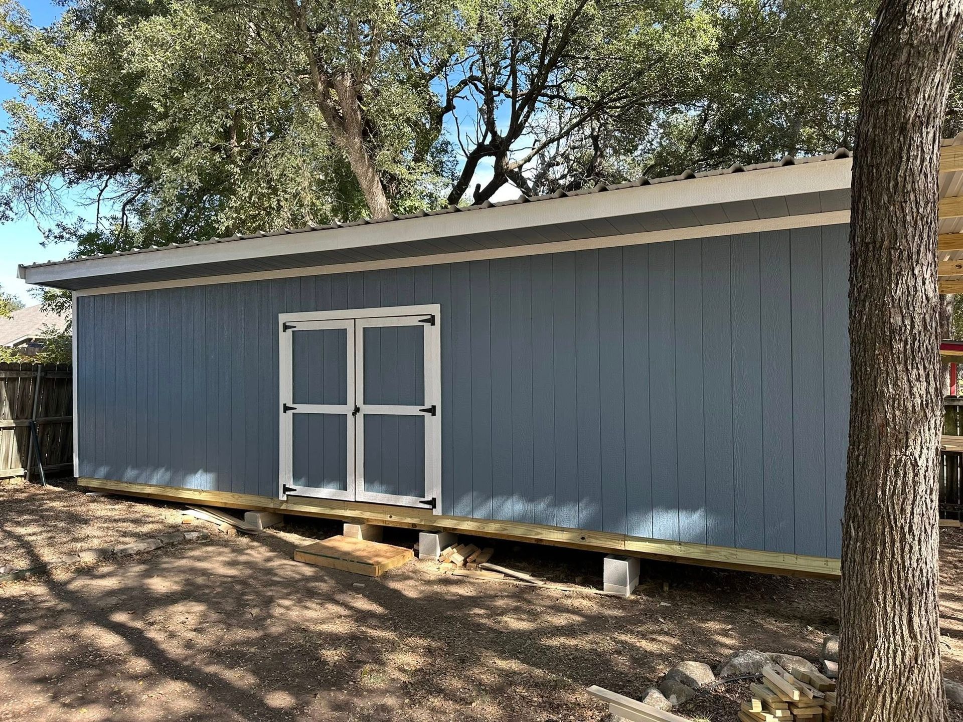 Blue shed with white double doors in a wooded yard, raised on blocks under large trees.