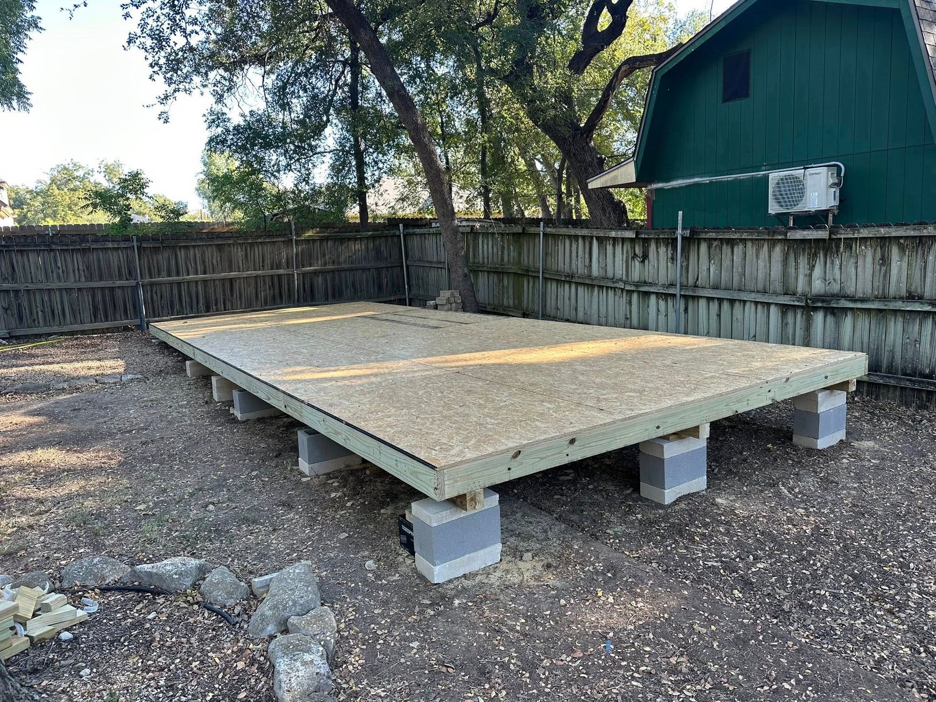 Raised wooden platform on cinder blocks in a fenced backyard, with trees and a green shed nearby