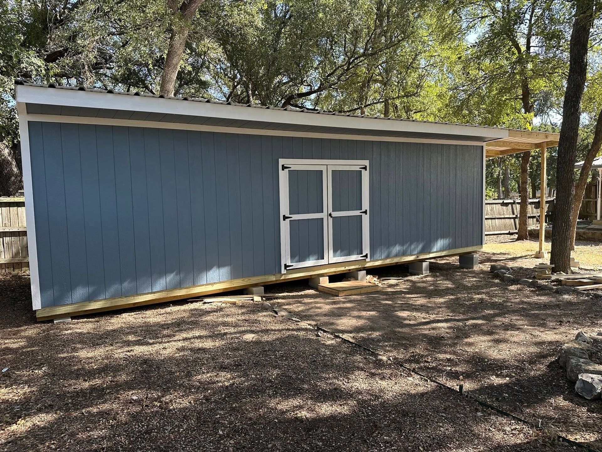 Blue shed with white double doors on a gravel lot, surrounded by trees.