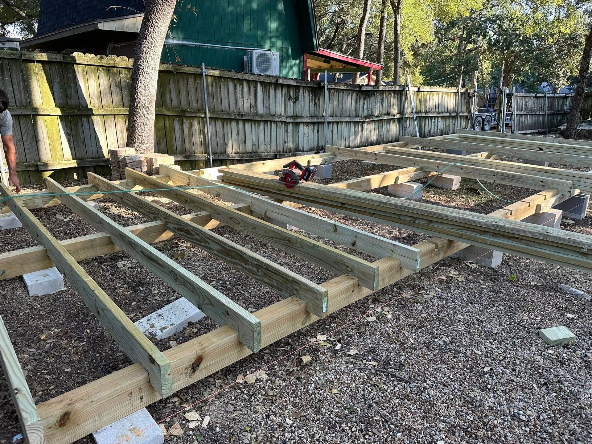 Wooden deck framing under construction in a backyard, with gravel ground and a fence in the background
