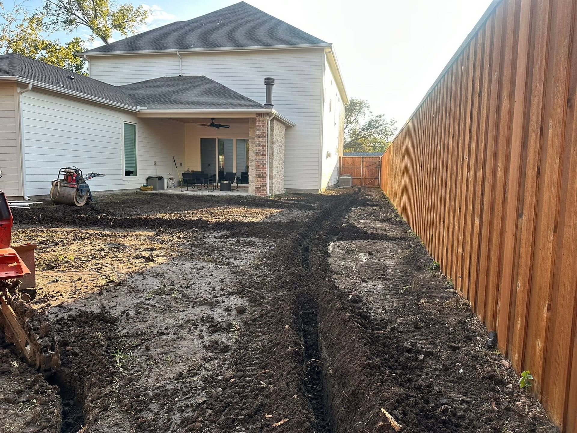Dug-up backyard beside a white house and tall wooden fence, with a trench and bare soil.