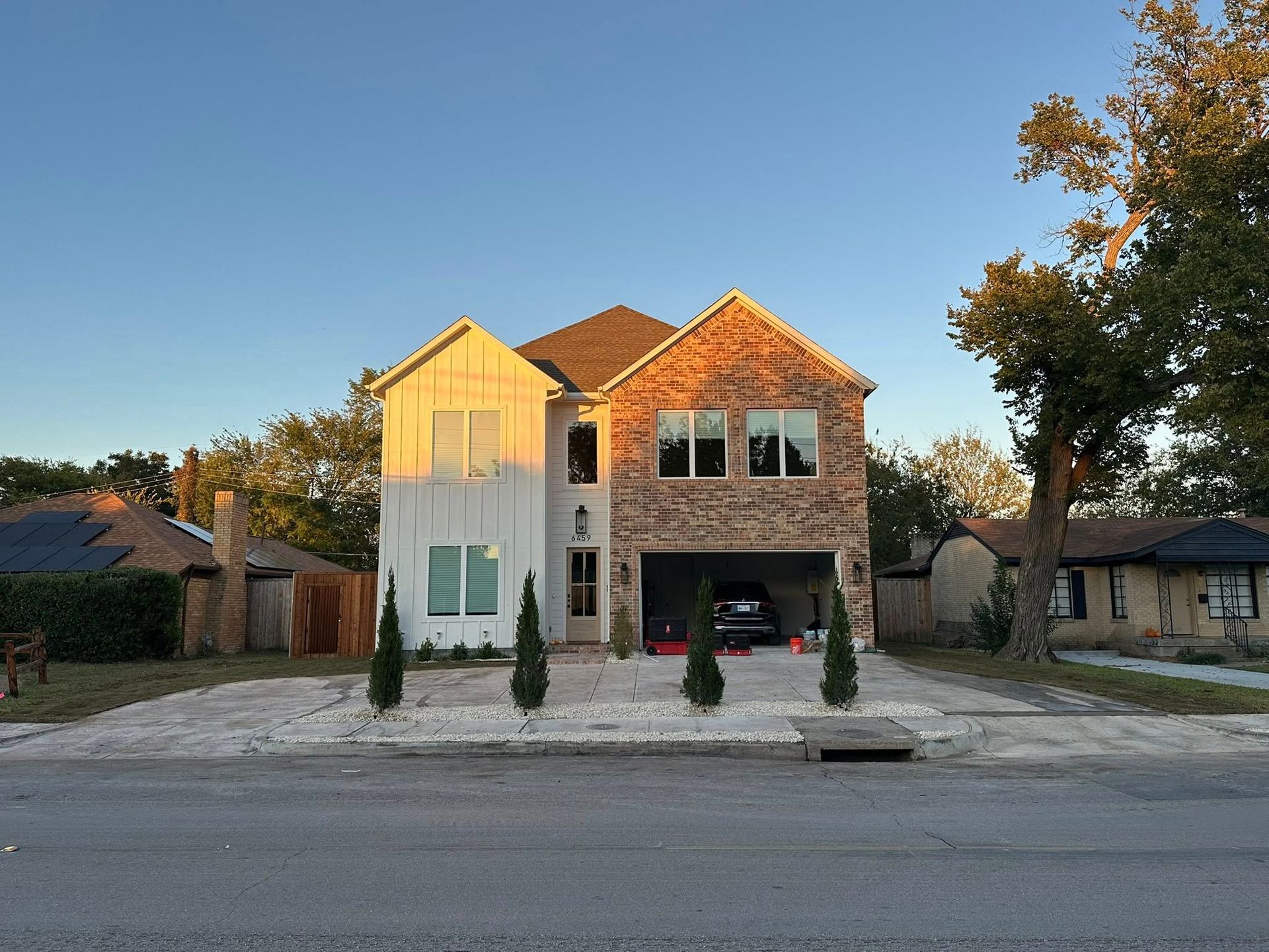 Modern two-story house with white and brick facade, trees, and a driveway at sunset