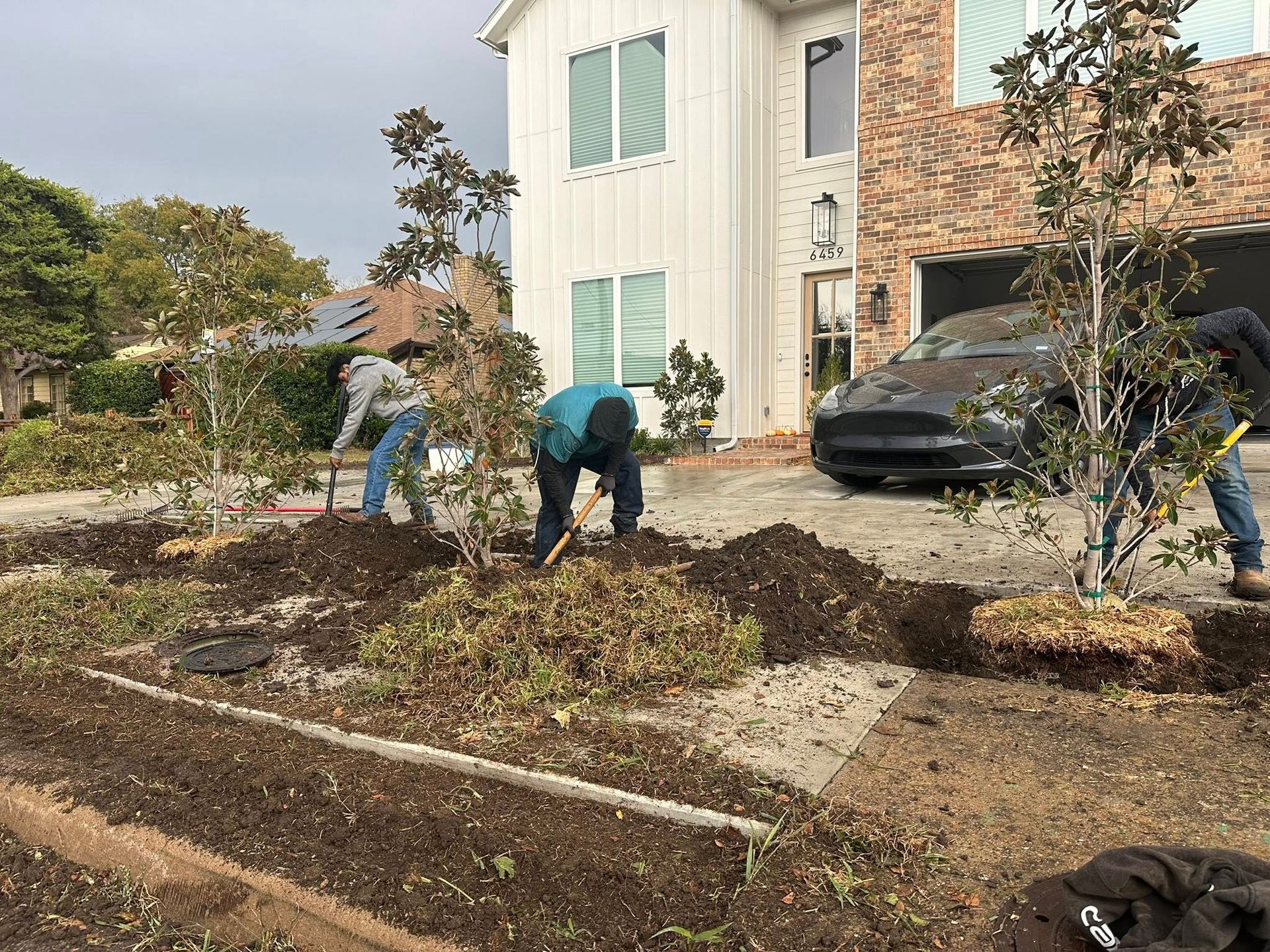 People gardening in front of a house, clearing a landscaped bed with a parked car nearby