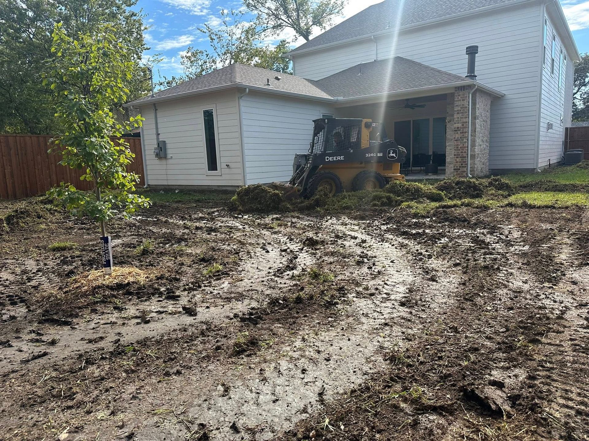 Mud-covered yard beside a white house with a patio and deck in bright sunlight