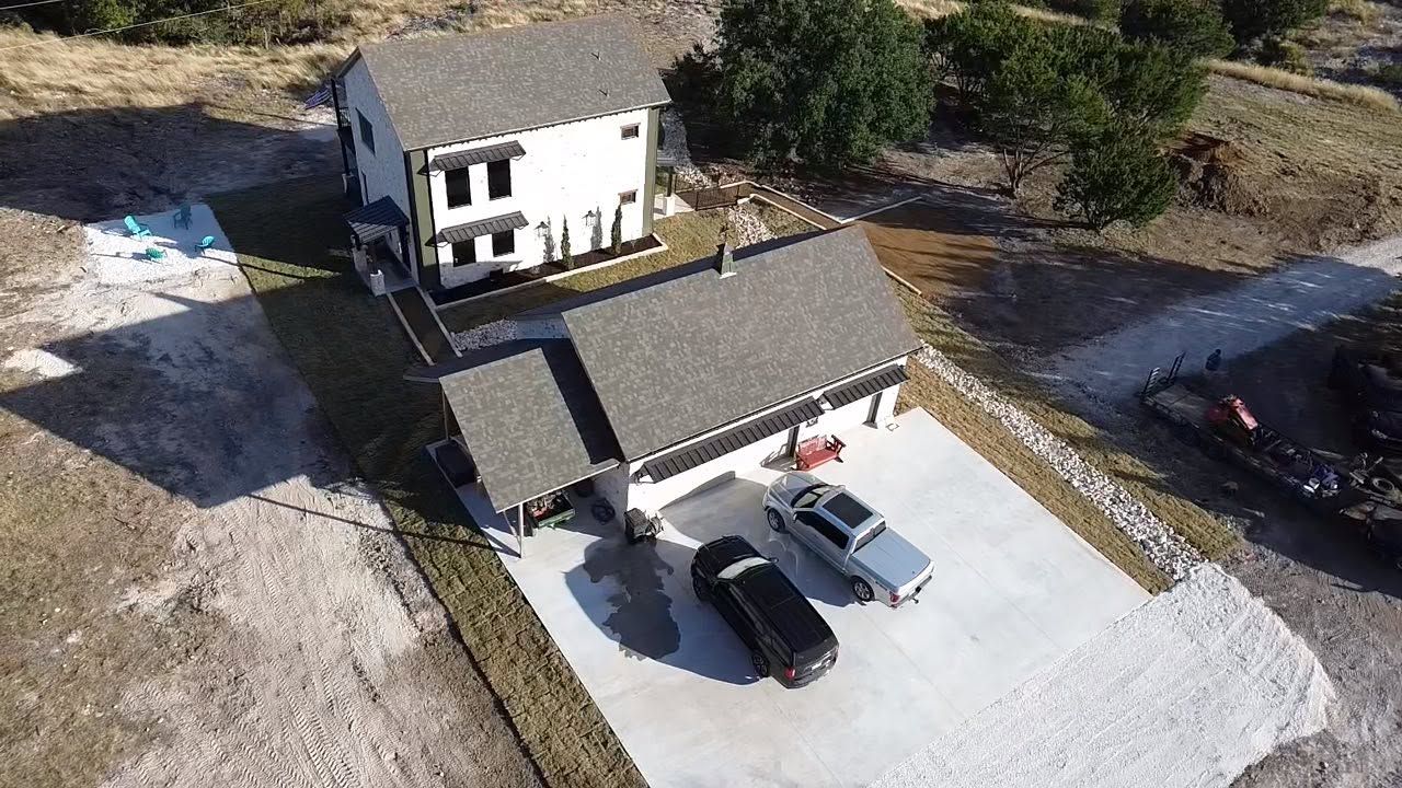 Aerial view of a white house with a dark roof, attached garage, and two cars parked on a gravel driveway.