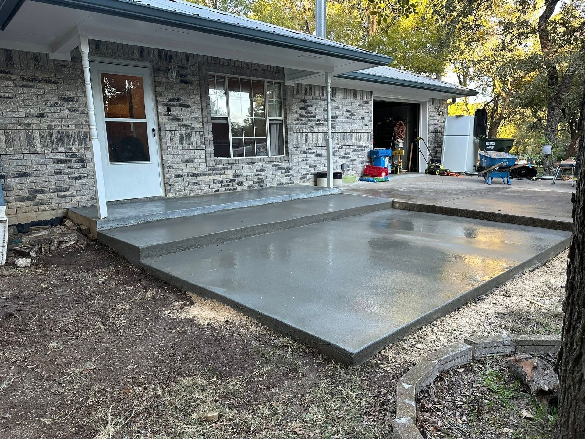 Freshly poured concrete patio slab beside a stone house with a white door and windows