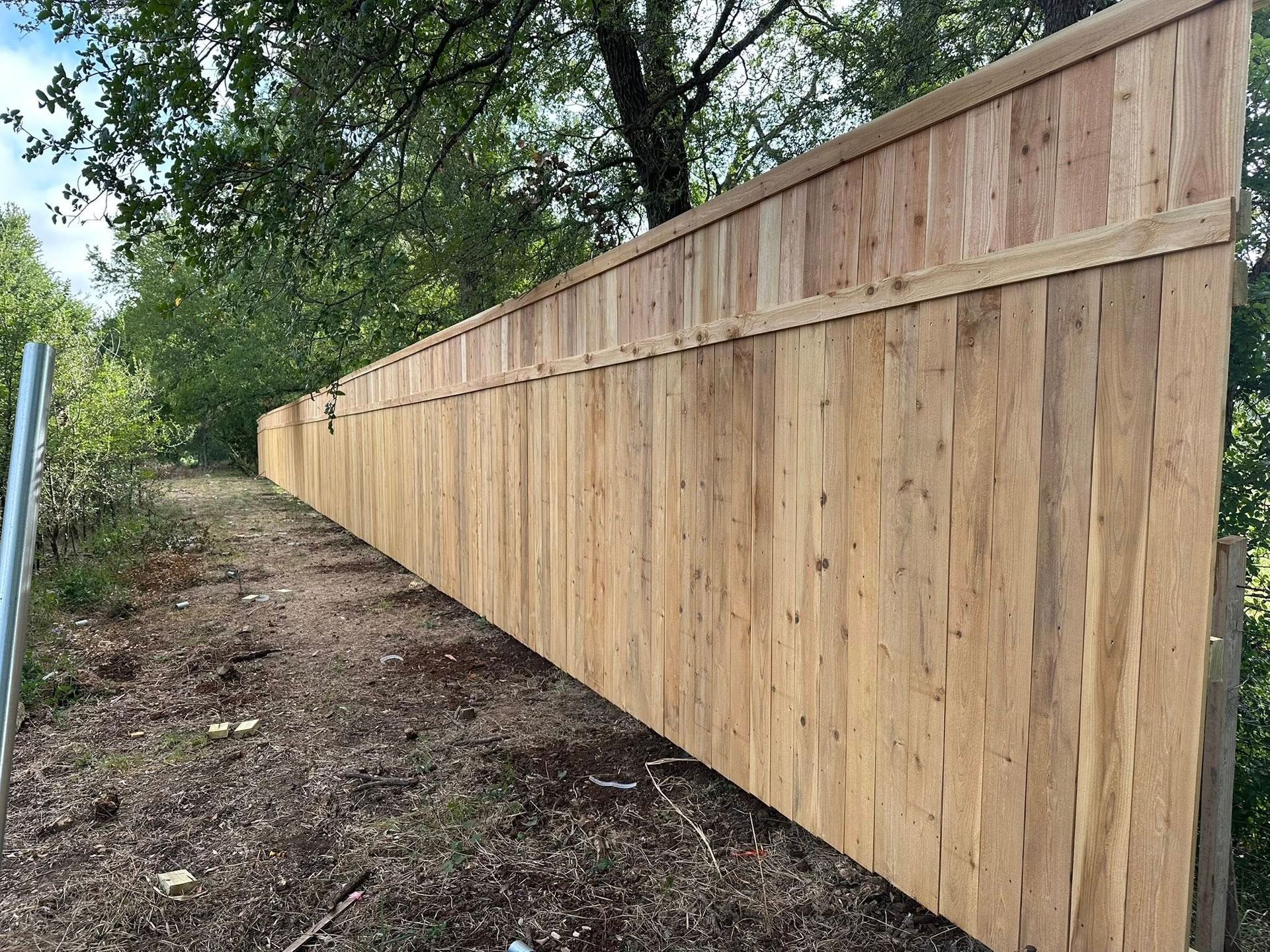Long wooden privacy fence along a dirt path beside trees and brush