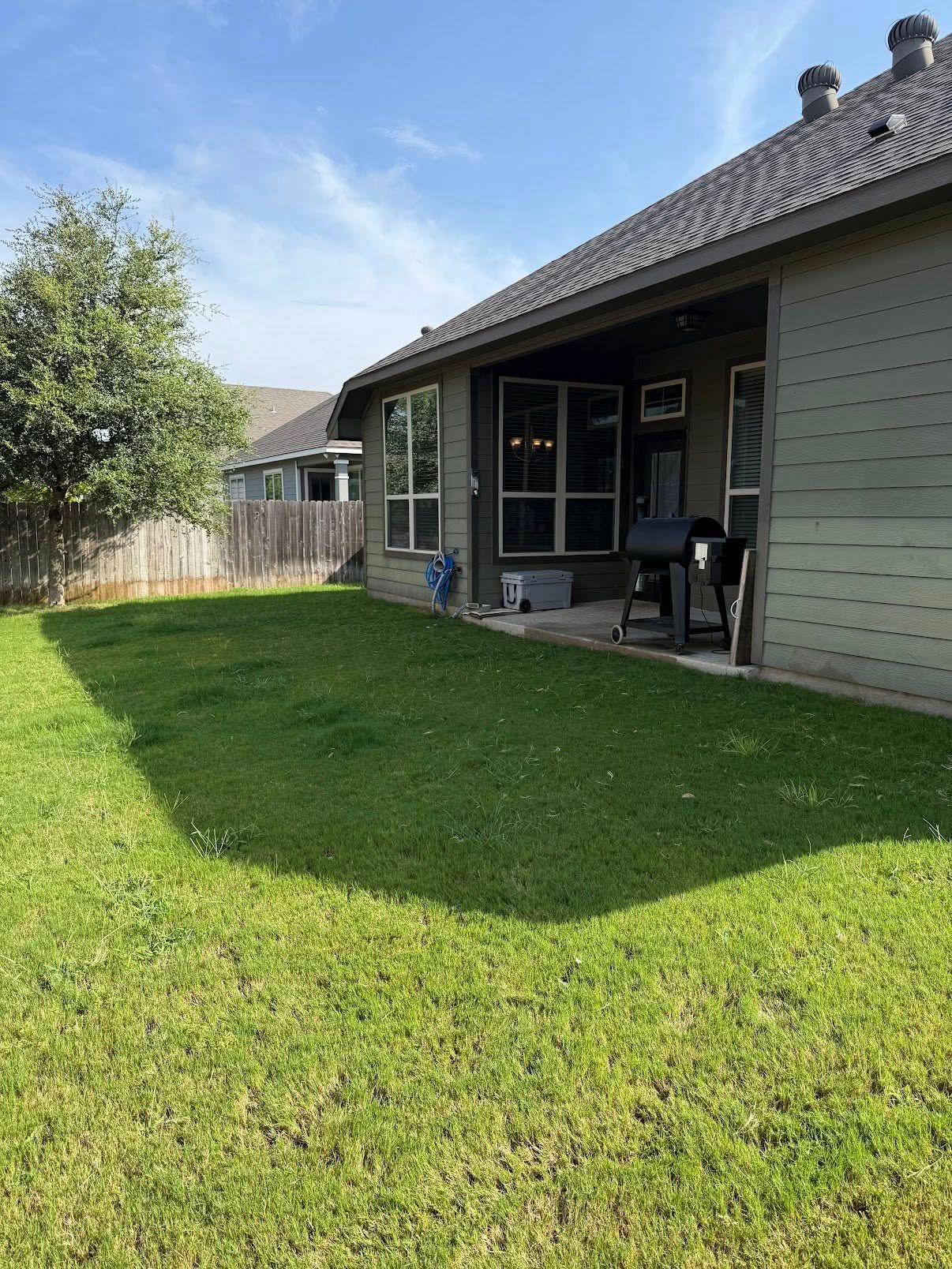 Backyard lawn beside a covered patio with a table and chairs, trees and a wooden fence nearby