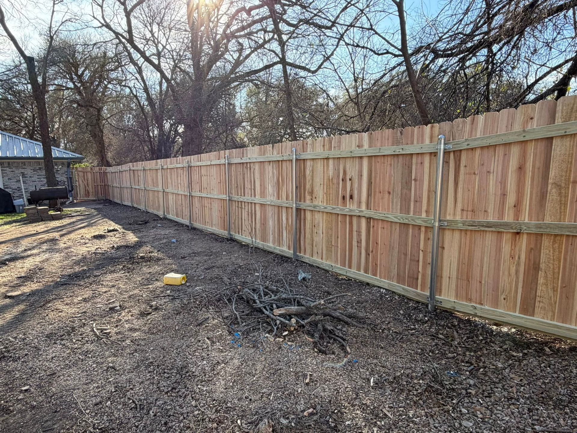Wooden privacy fence along a dirt yard with leafless trees and scattered debris near a house