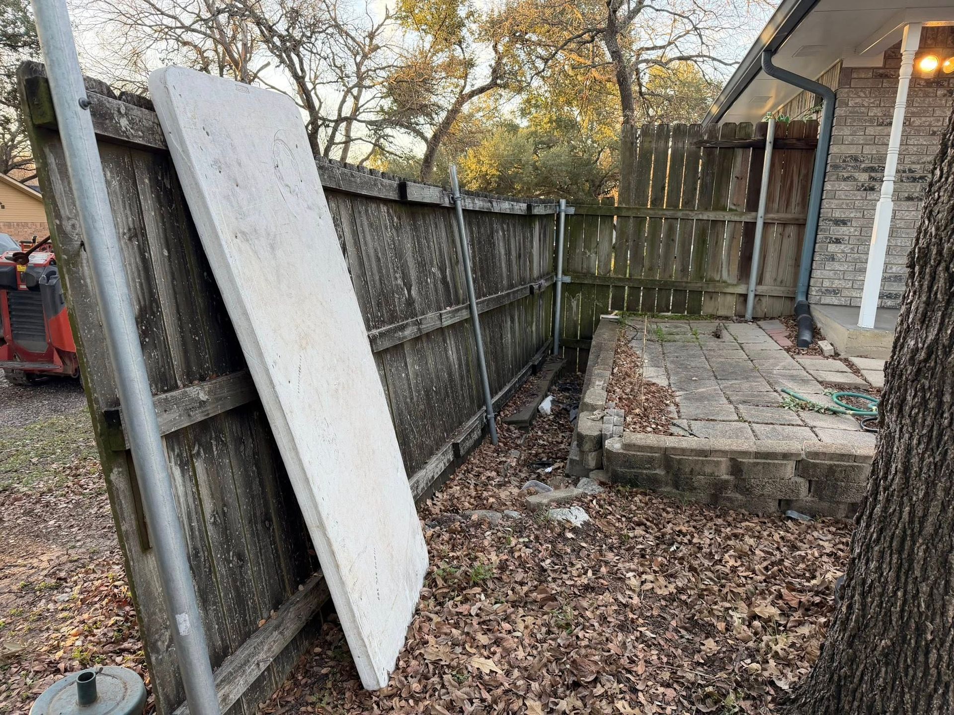 Large white board leaning against a wooden fence beside a small brick patio with fallen leaves.