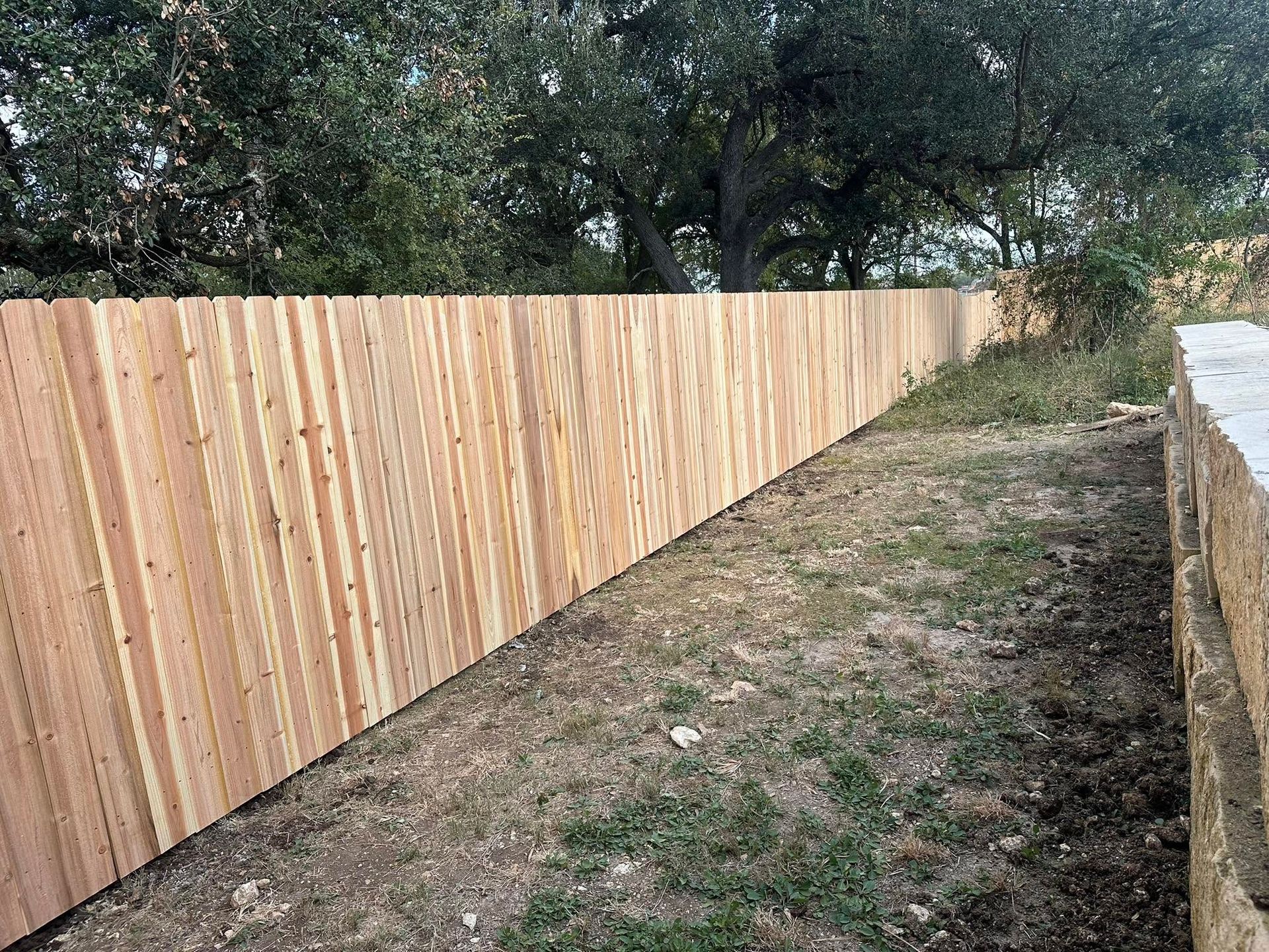 Wooden fence lines a narrow grassy path beside trees and a white wall.