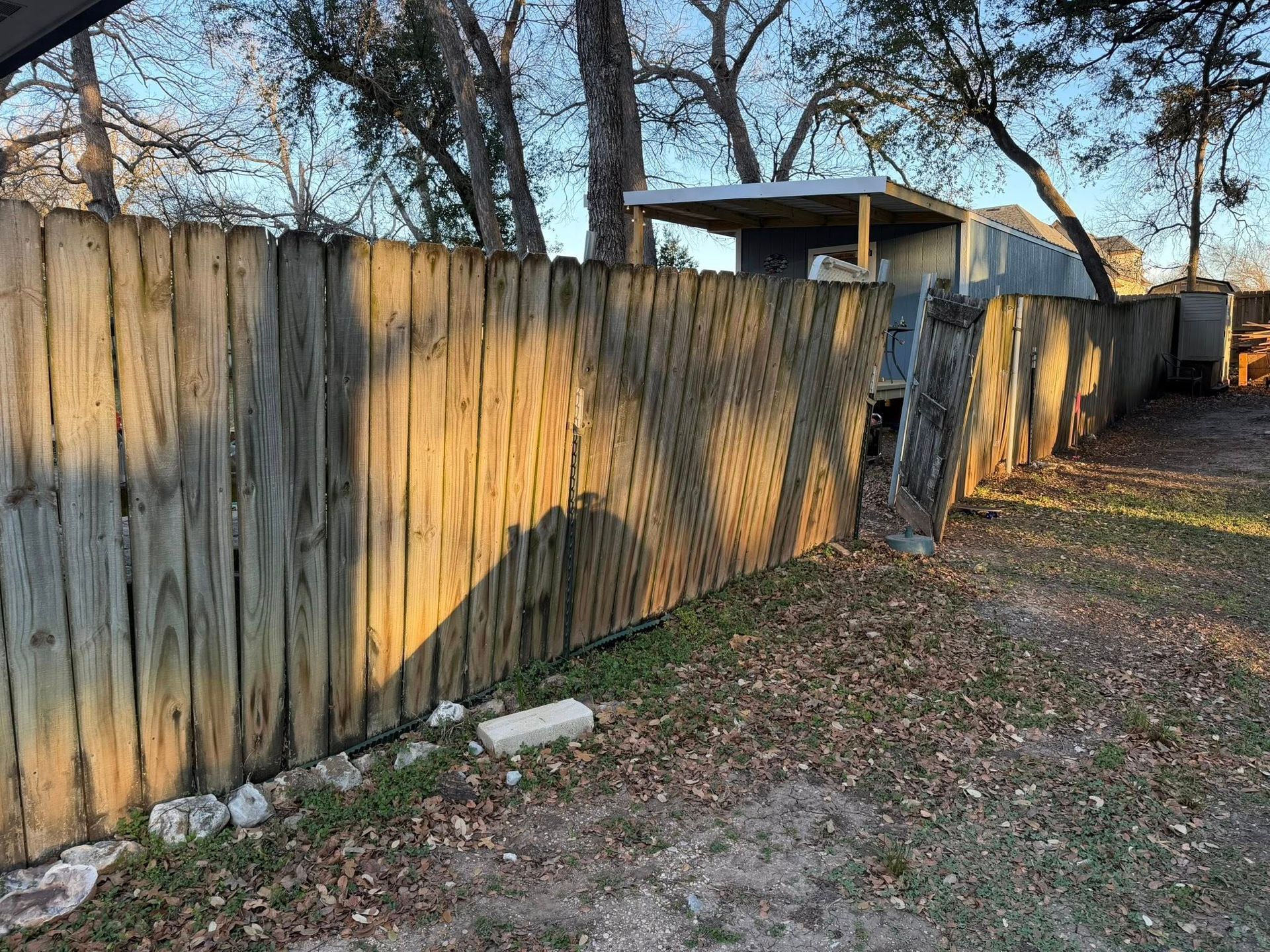 Weathered wooden fence along a dirt path with trees and a small shed in the background