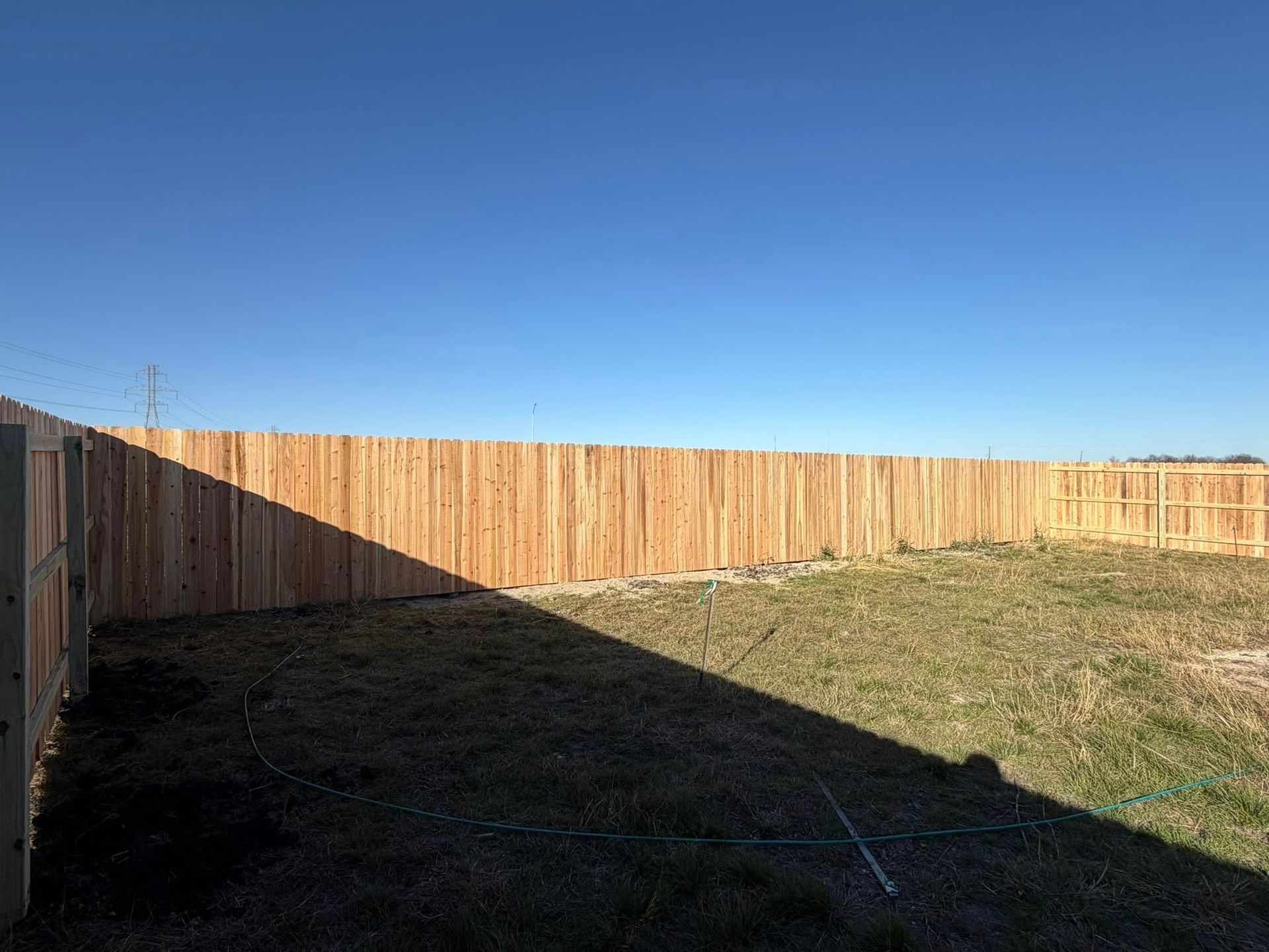 Backyard with a wooden fence, blue sky, and a large shadow over grass and dirt.