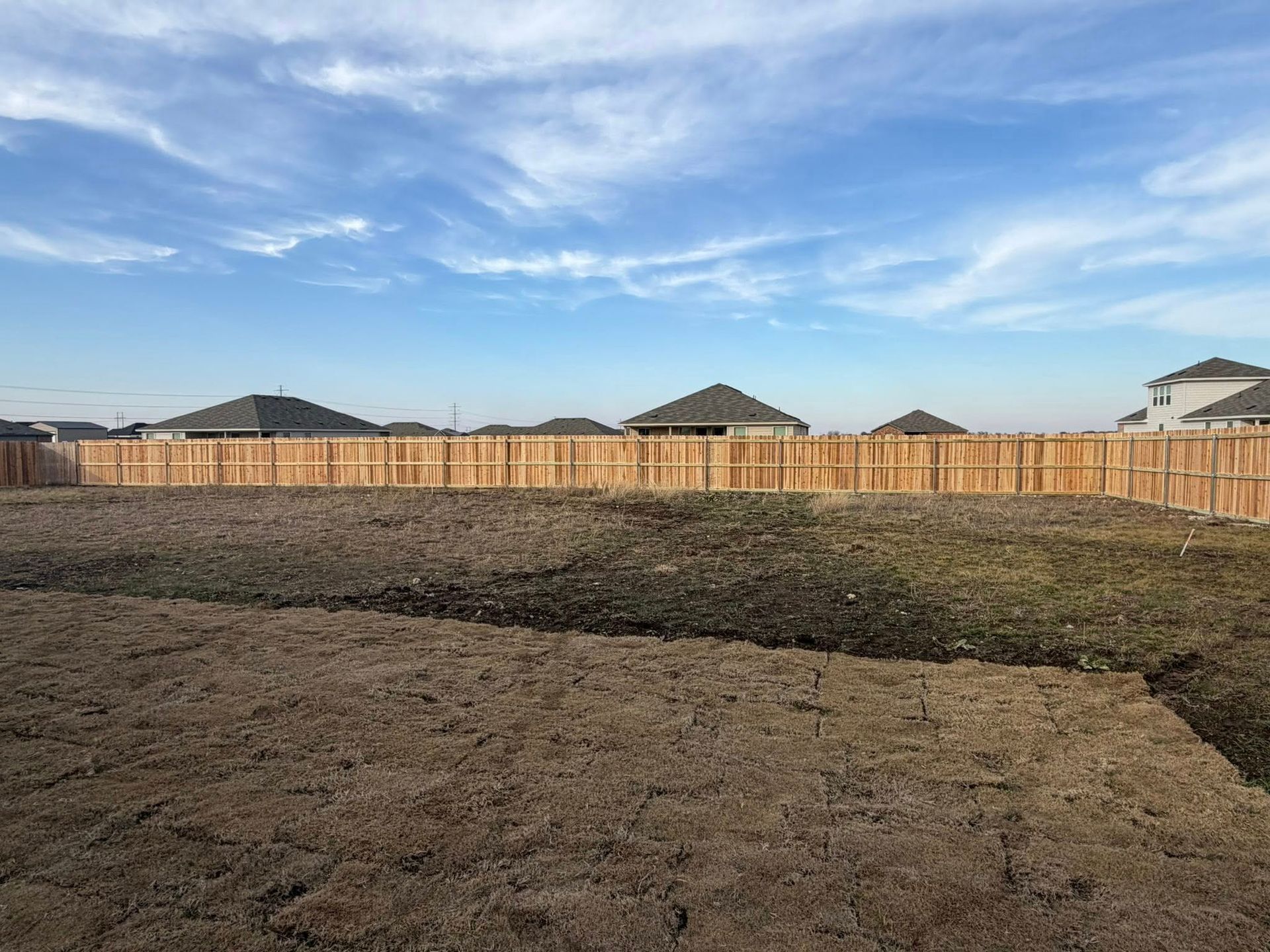 Vacant dirt lot beside a wooden fence under a blue sky with scattered clouds