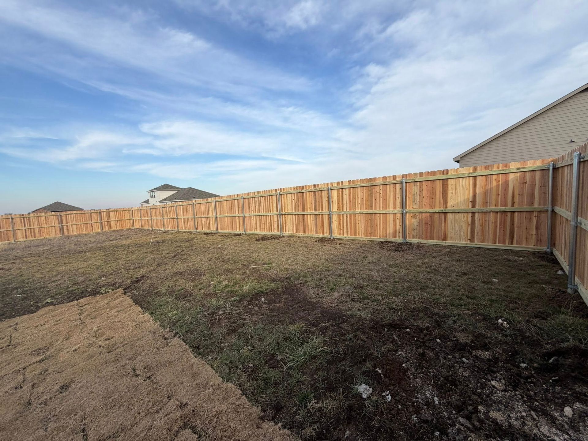 Empty backyard with a wooden fence, patchy dirt, and a blue cloudy sky