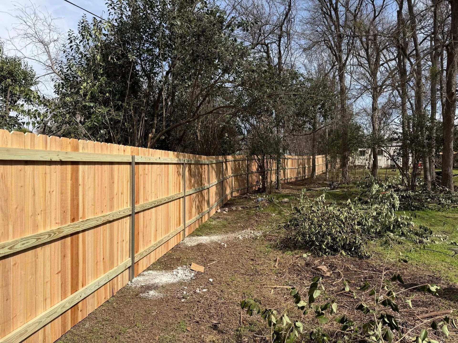 Wooden privacy fence along a dirt path beside trees and brush on a sunny day