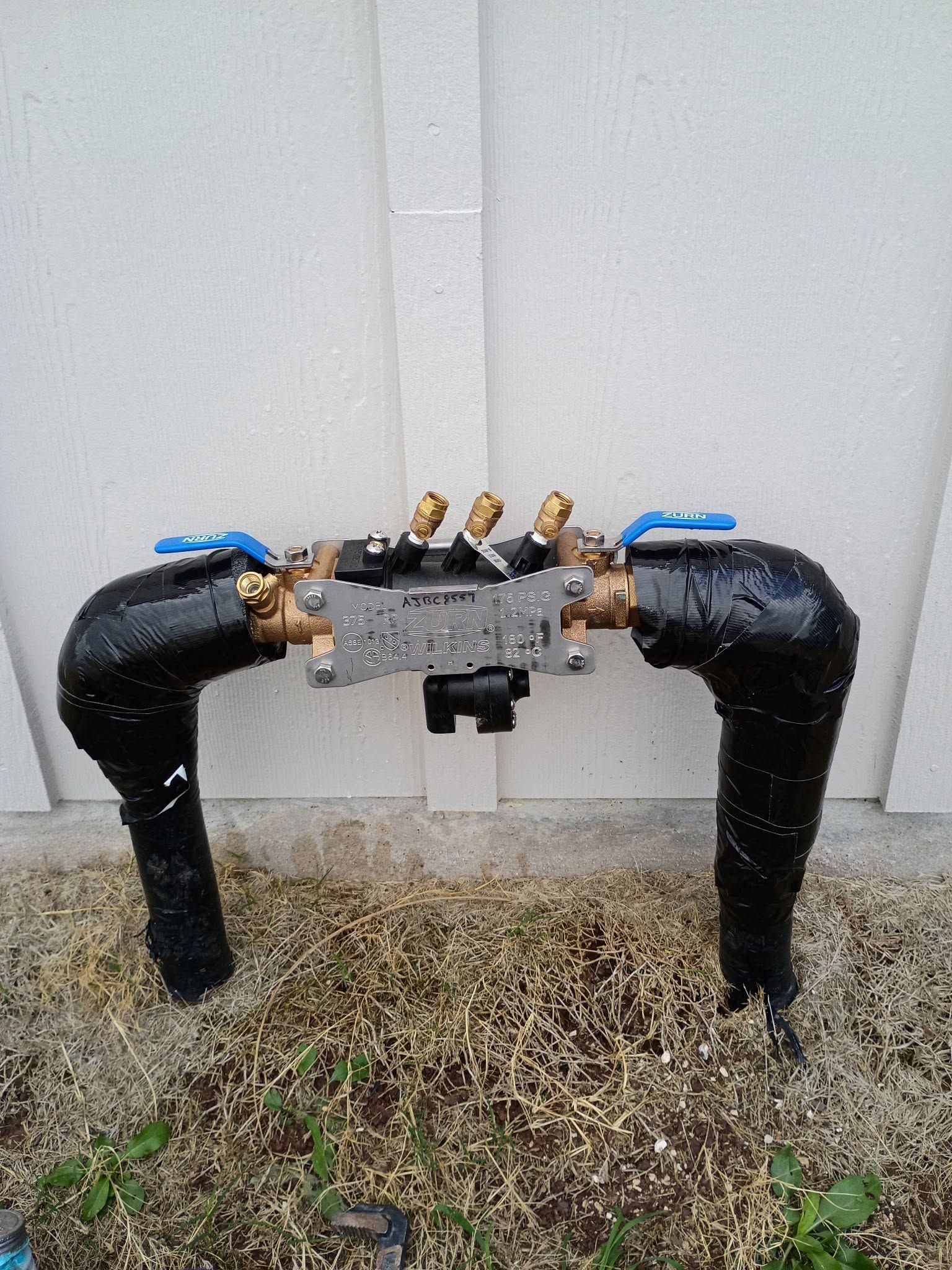 Outdoor black pipes with blue valves and brass fittings against a white wall.