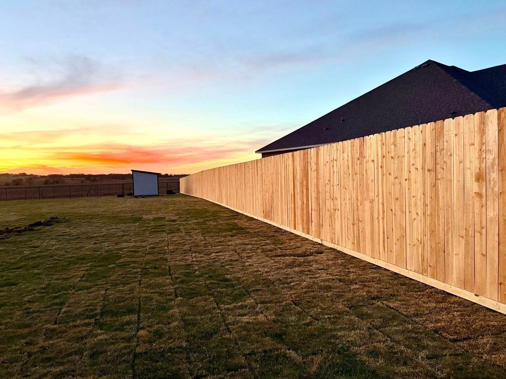 Long wooden fence beside a grassy yard at sunset, with a dark-roofed house and small shed in the distance.