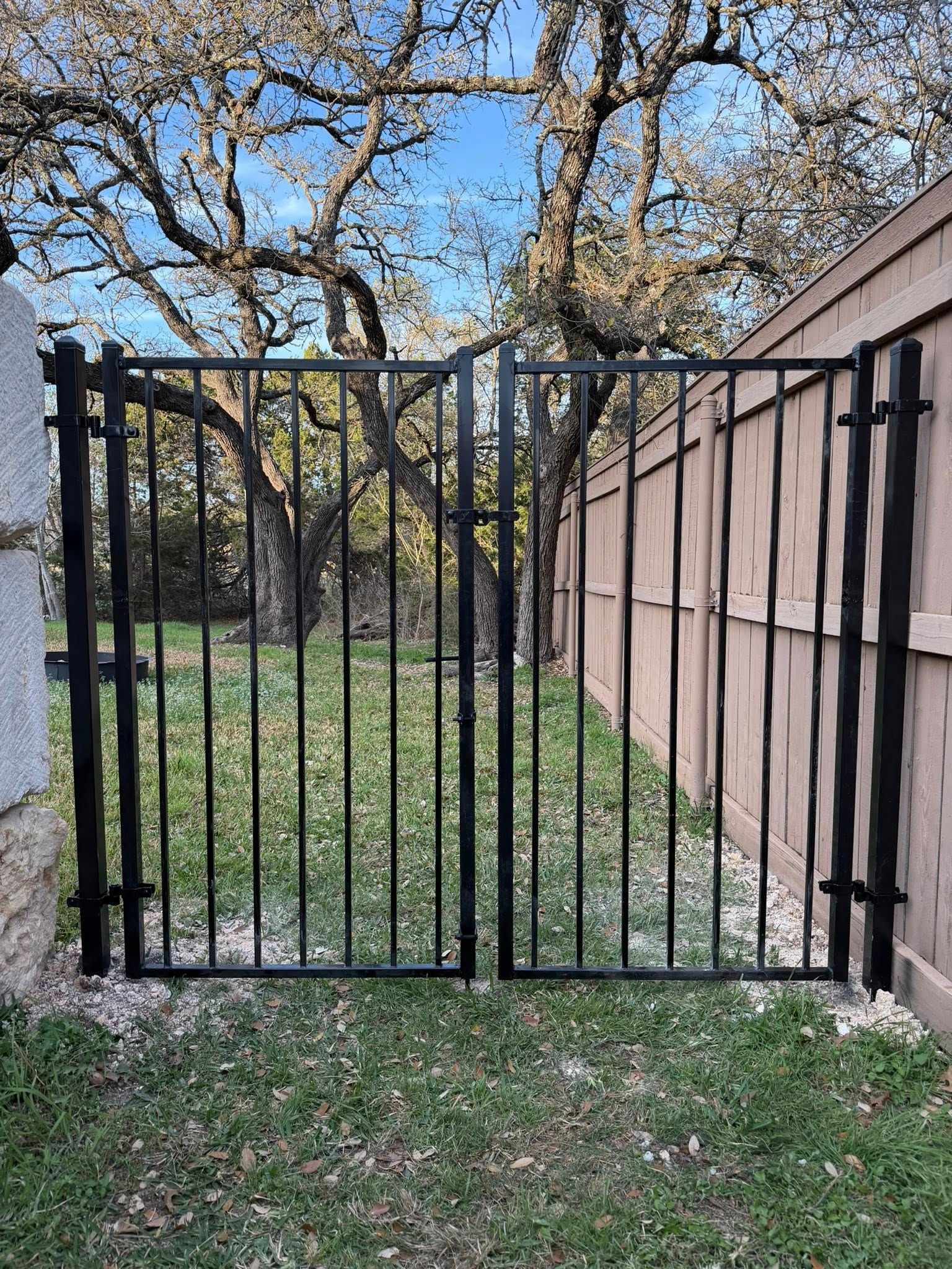Black metal gate between stone wall and wooden fence, opening to a grassy yard with leafless trees.