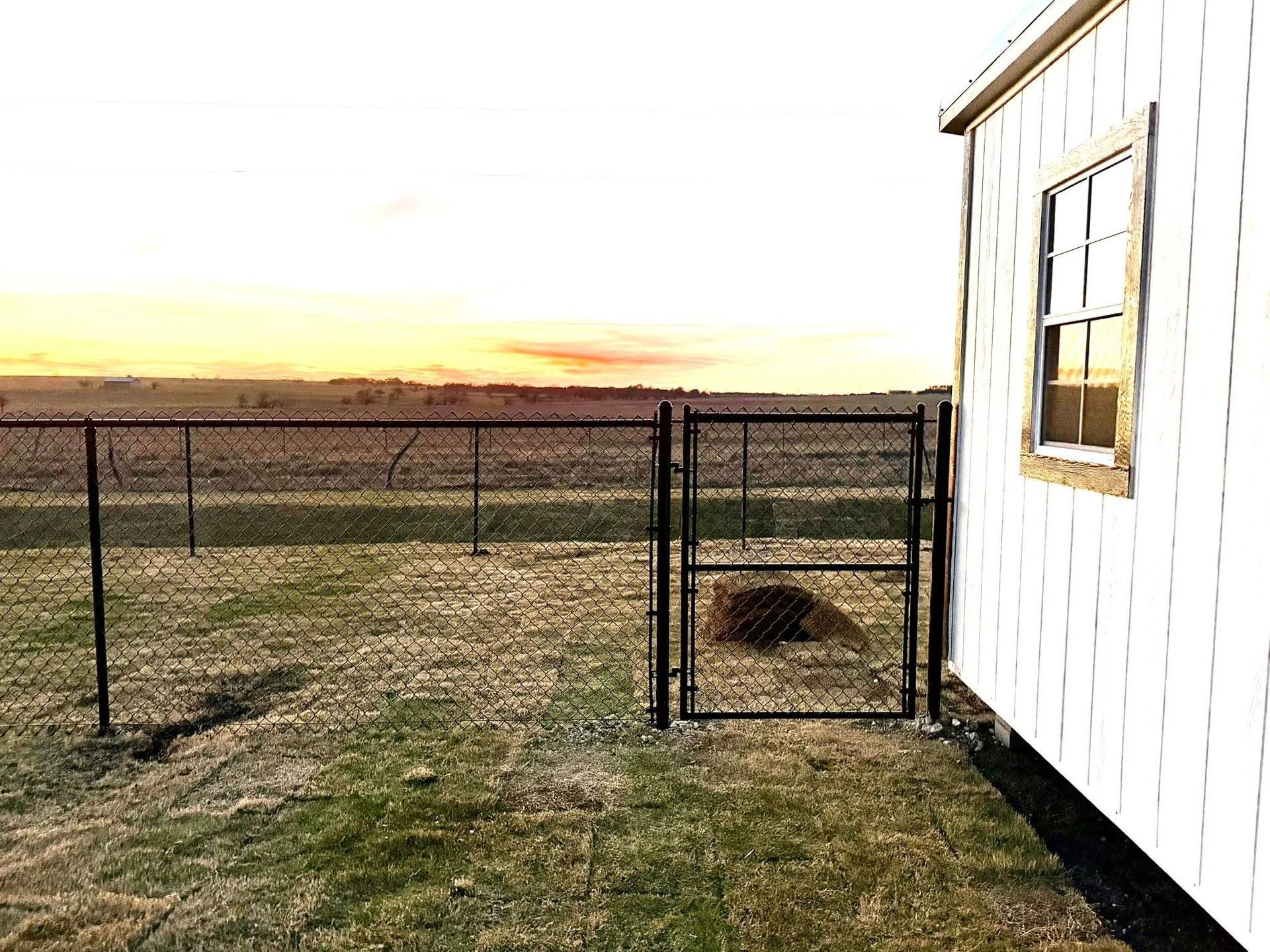 Chain-link fence and open gate beside a white building at sunset, with a field beyond.