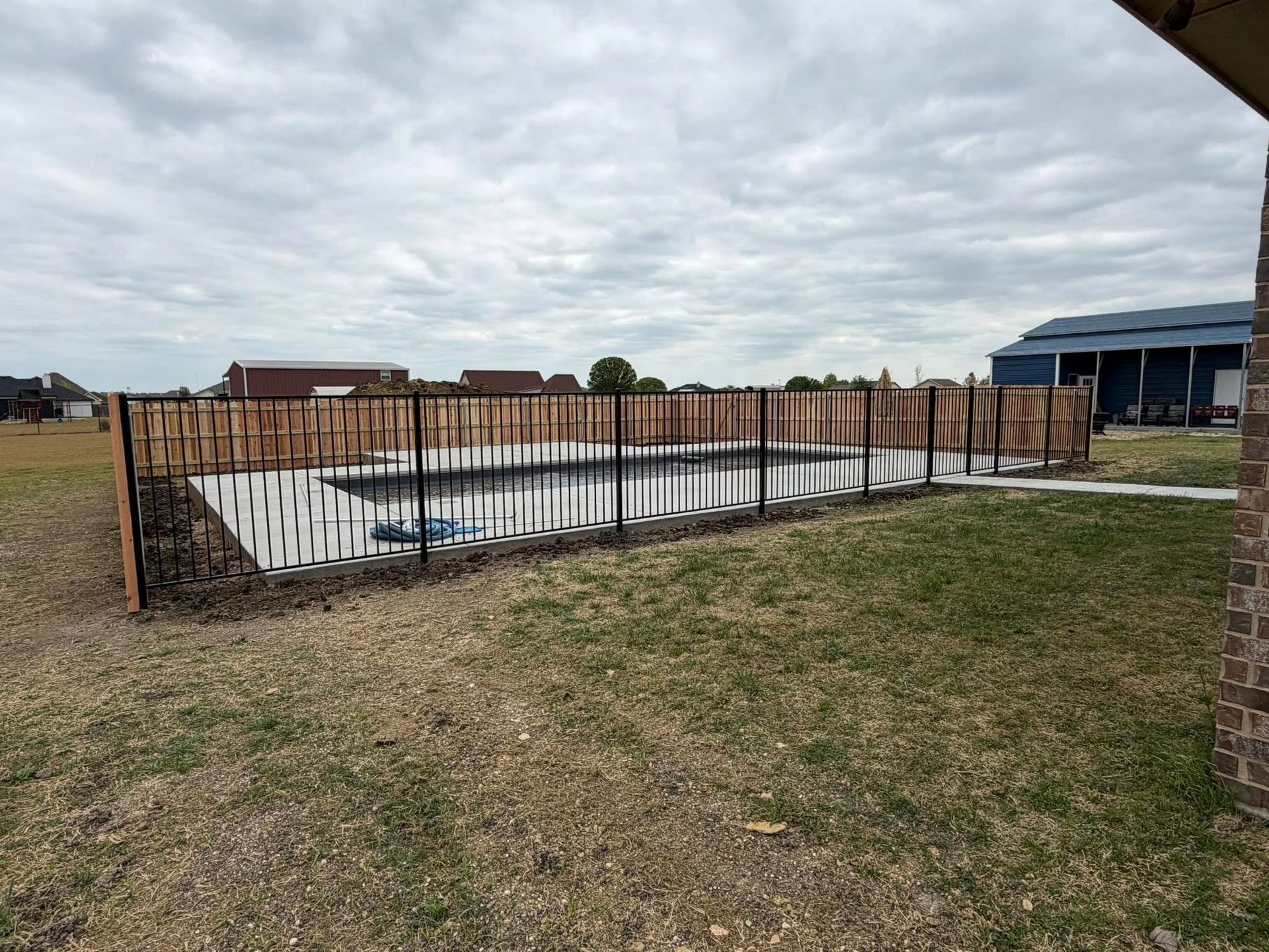 Backyard fenced-in inground pool under construction beside a grassy yard and brick house