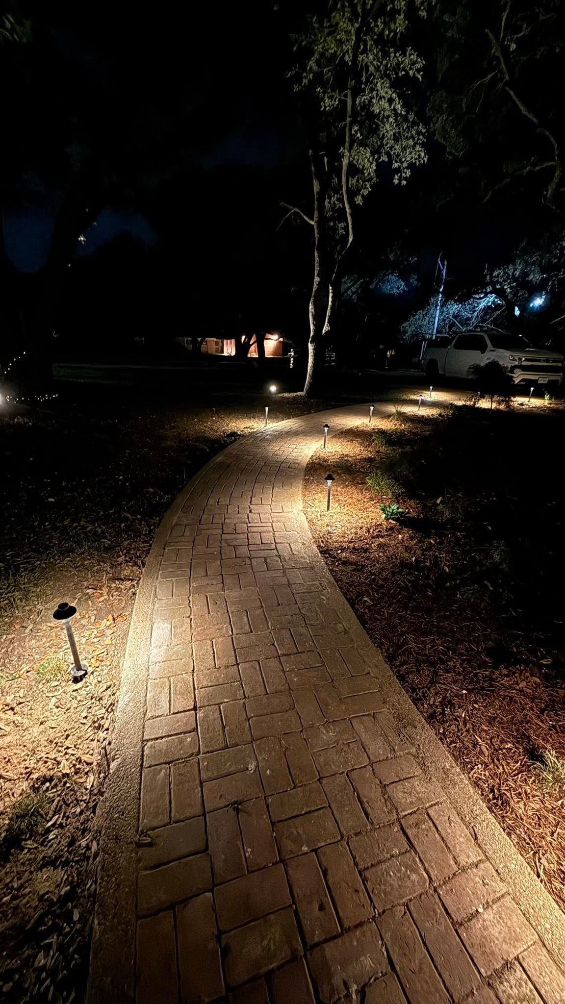 Dimly lit brick walkway through a park at night, lined with trees and scattered lights.