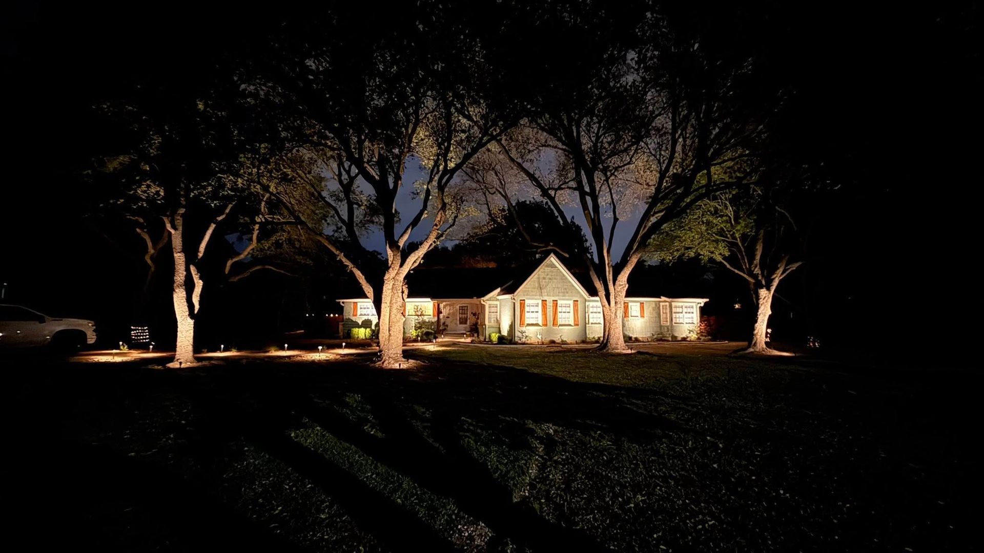 A lit house at night with trees in front and dark lawn shadows.