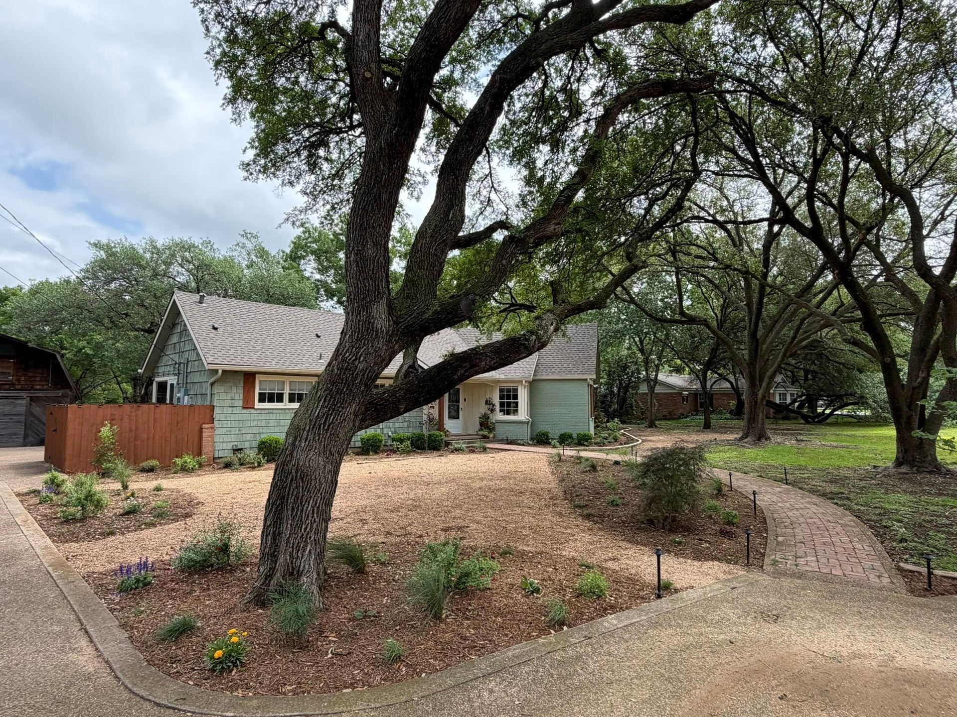 Tree-shaded front yard with gravel paths leading to a small house and landscaped garden beds