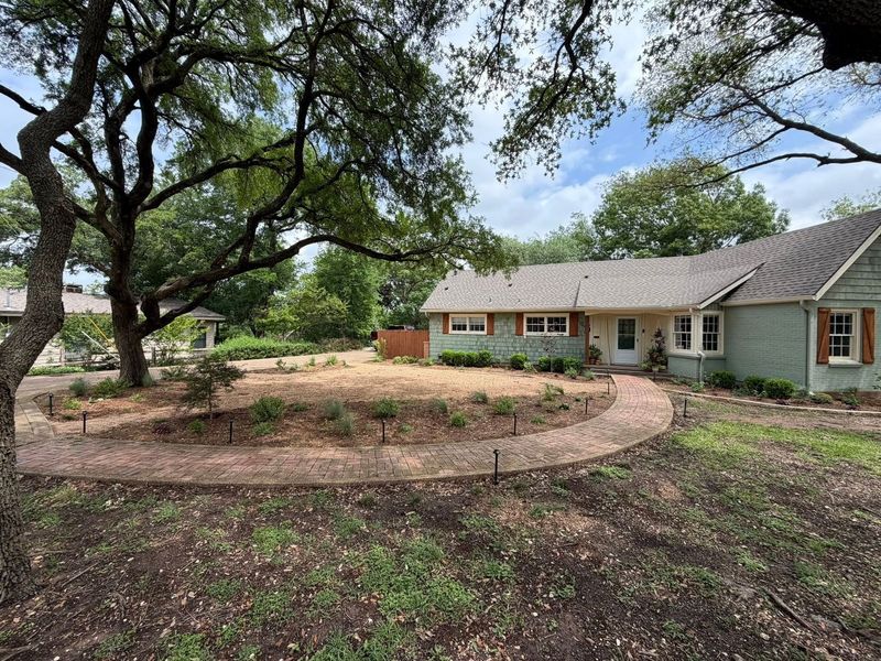 Single-story house with a curved stone path, large trees, and a dry front yard