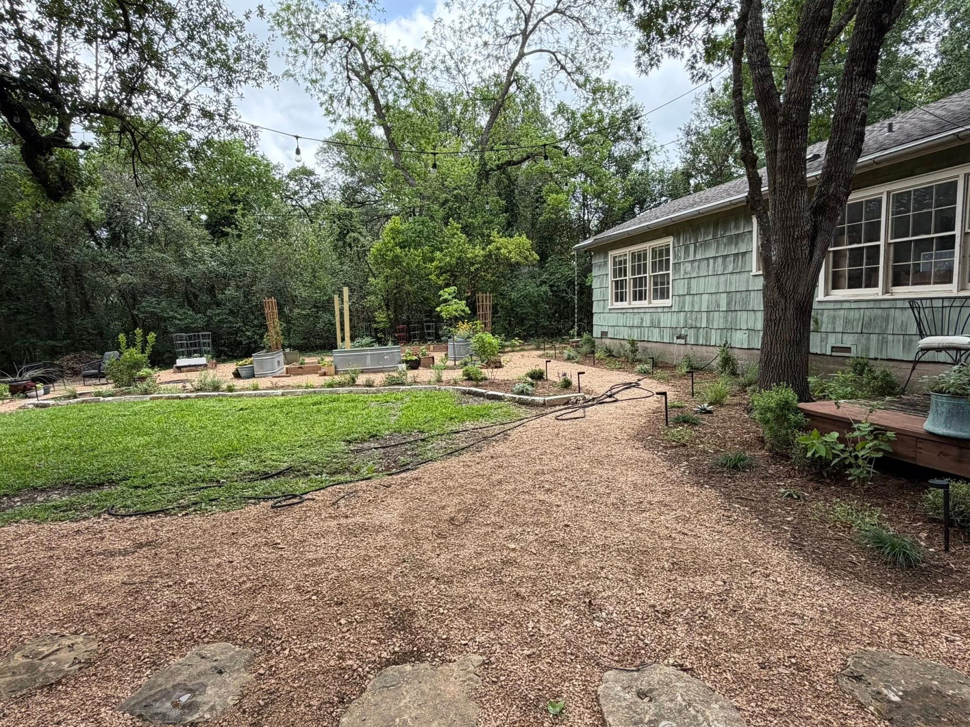 Wood-chip garden path beside a house, with trees, flower beds, and a small pond area.