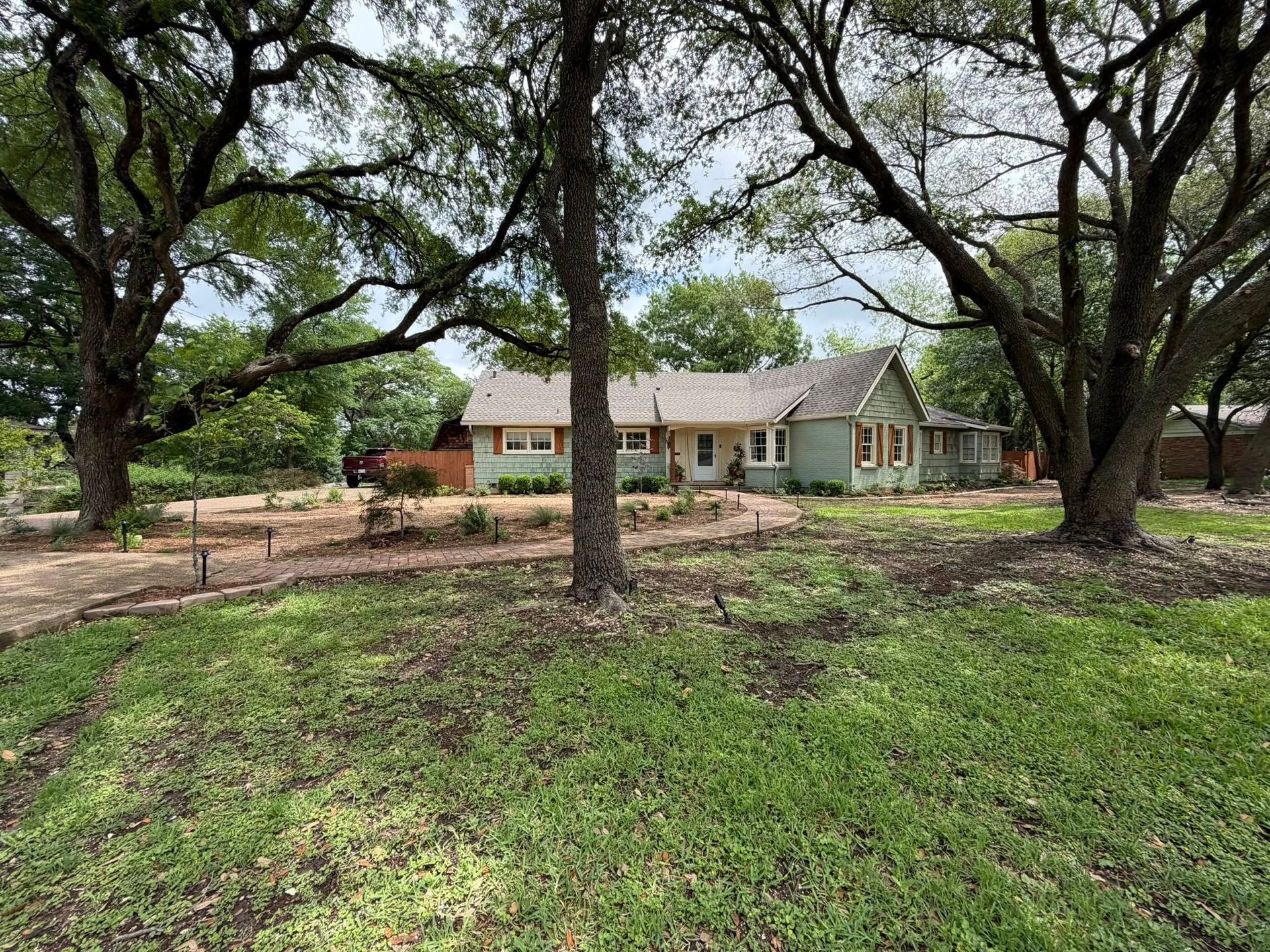 Single-story house with green shutters behind large oak trees and a grassy yard