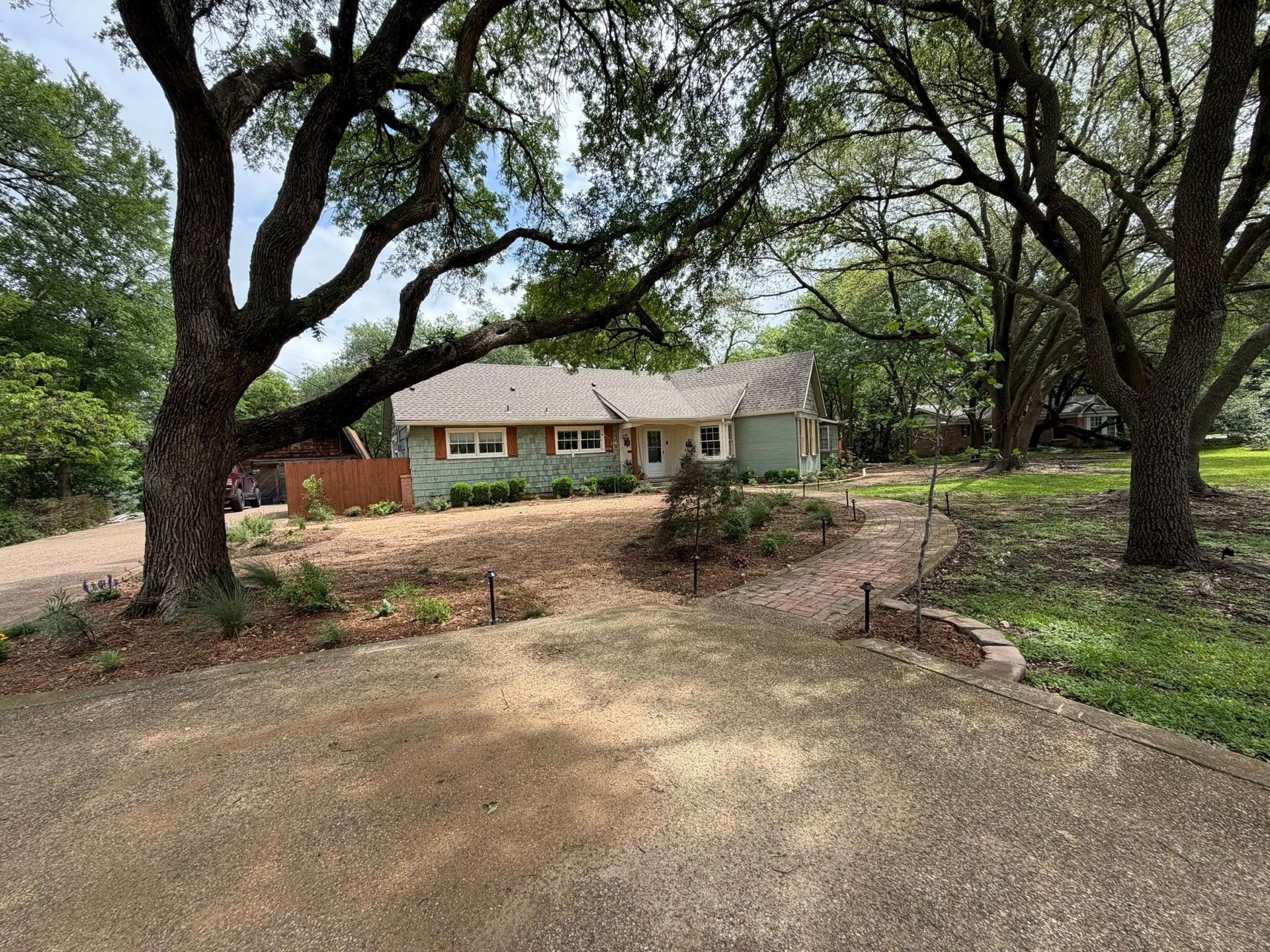Single-story house with light brick exterior, framed by large oak trees and a dirt driveway.