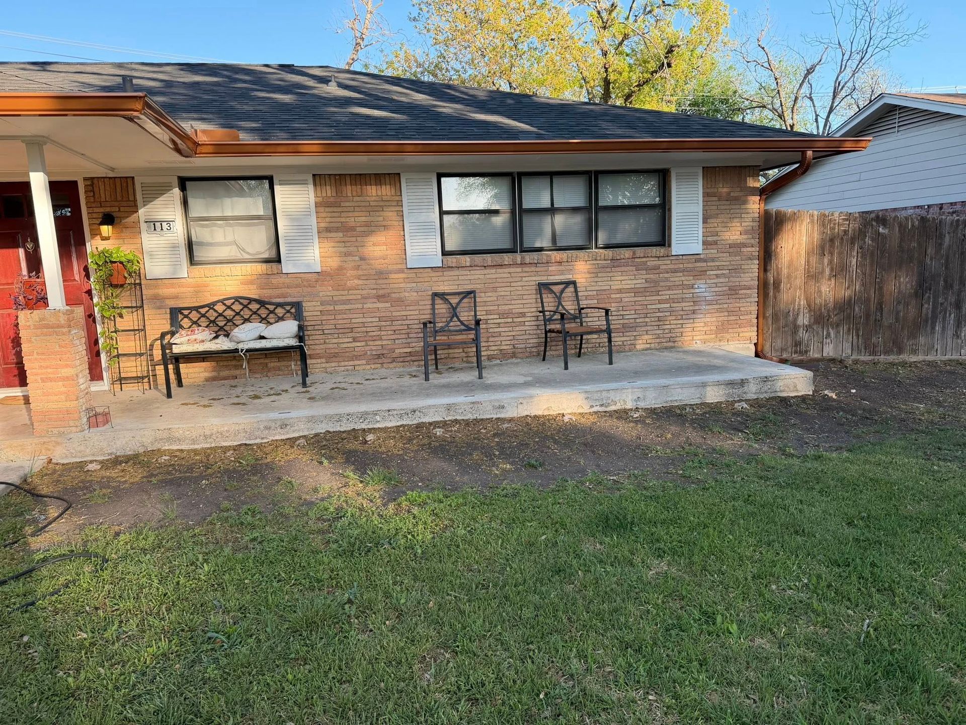 Brick house front porch with two chairs, a bench, and a grassy yard