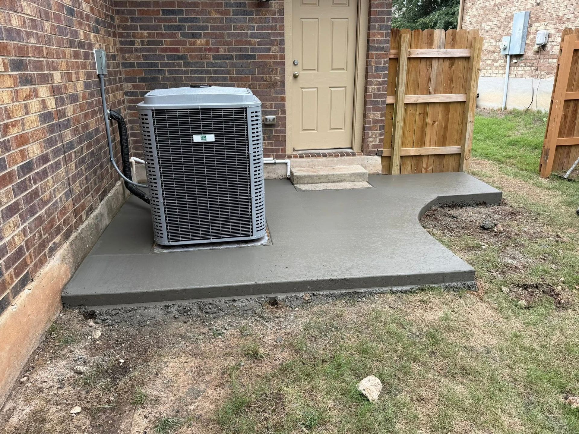 Concrete patio beside brick house with HVAC unit, door, and wooden fence.