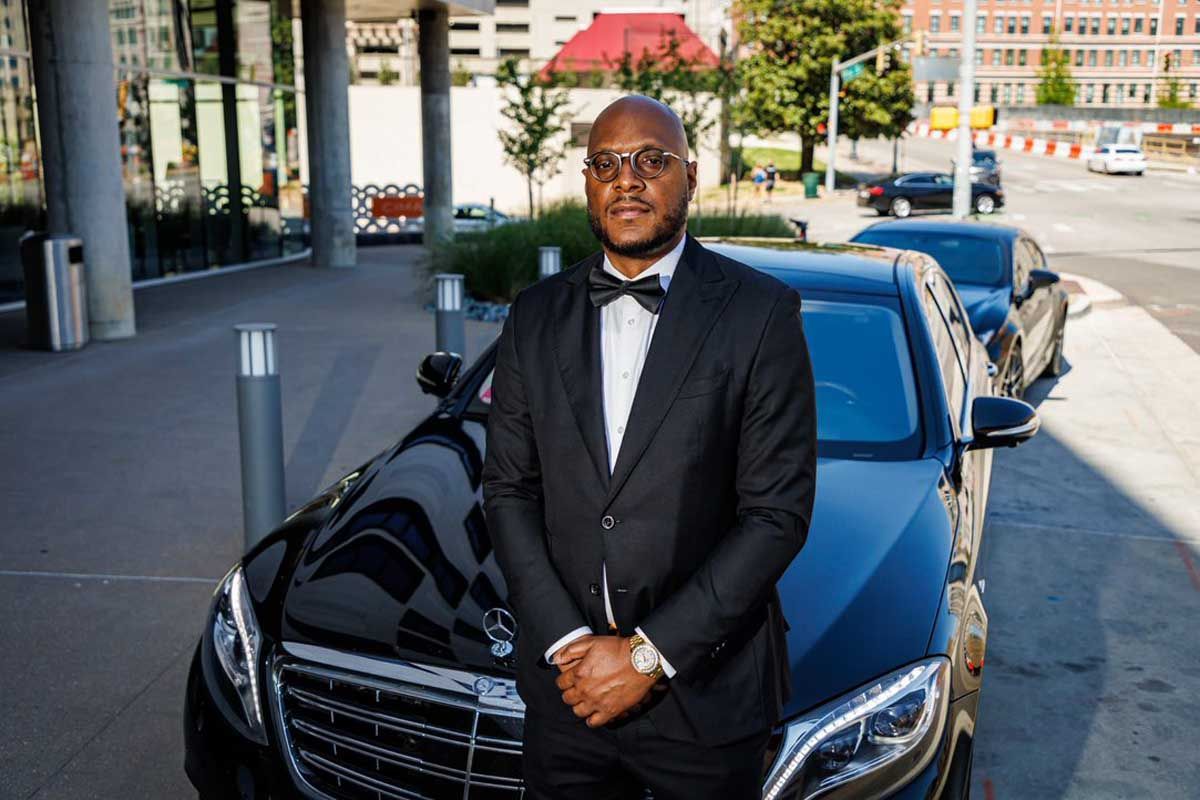 A professional chauffeur in black suit and bow tie stands in front of a black car. True Concierge