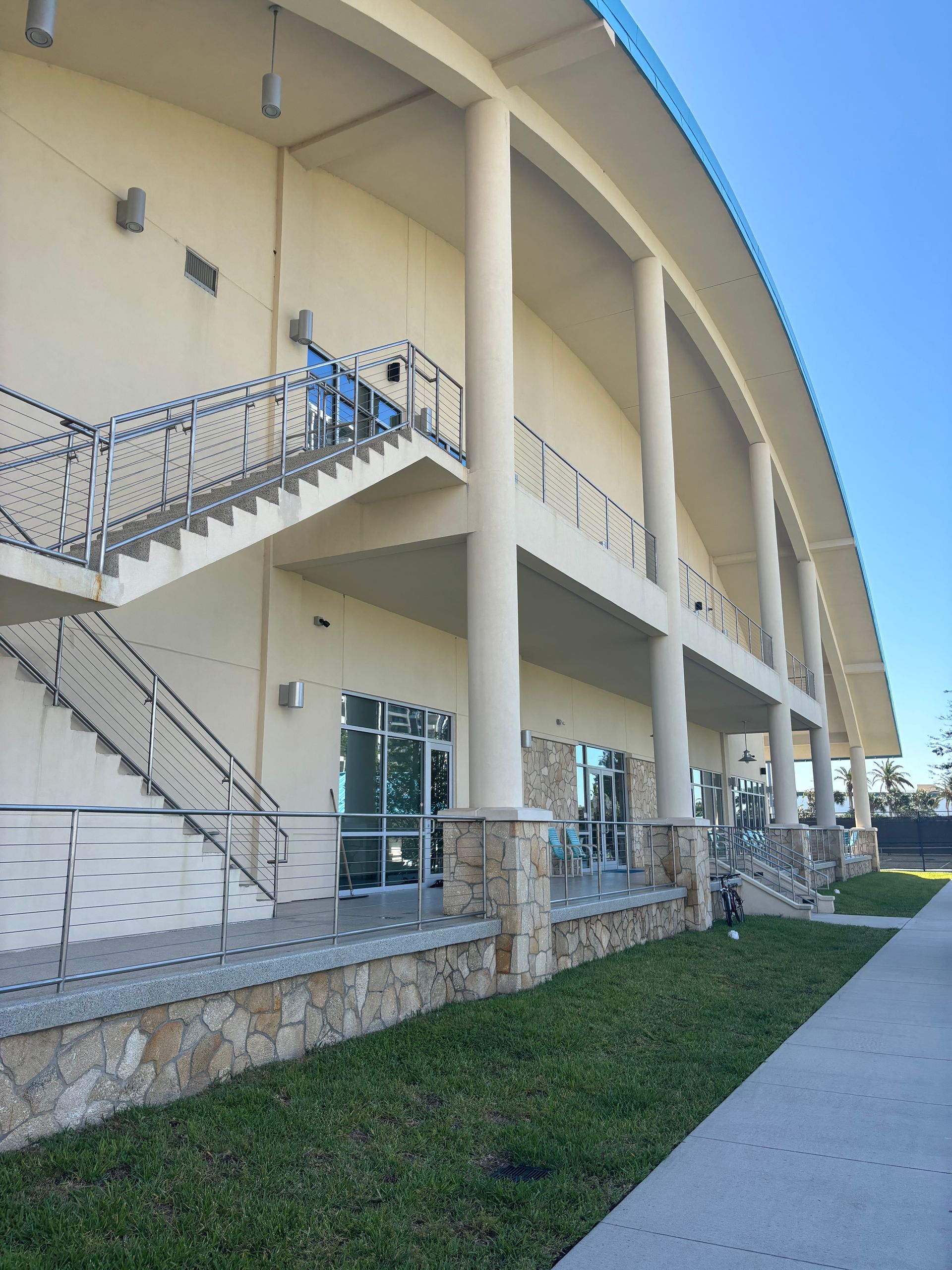 Exterior of a beige building with stairs, railings, and a grassy walkway under a bright blue sky