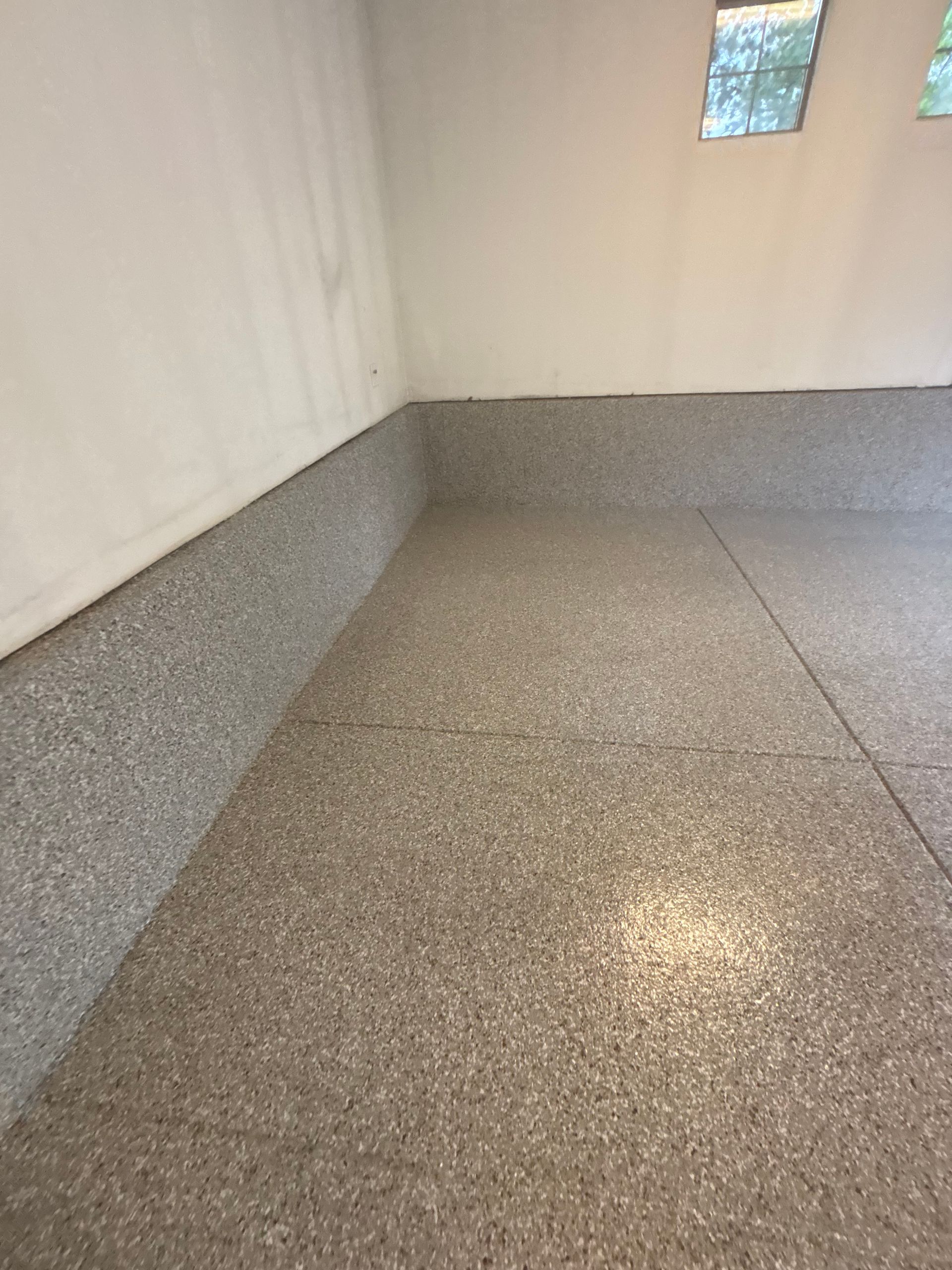 Empty room with white walls and a gray speckled floor, lit by a small window.