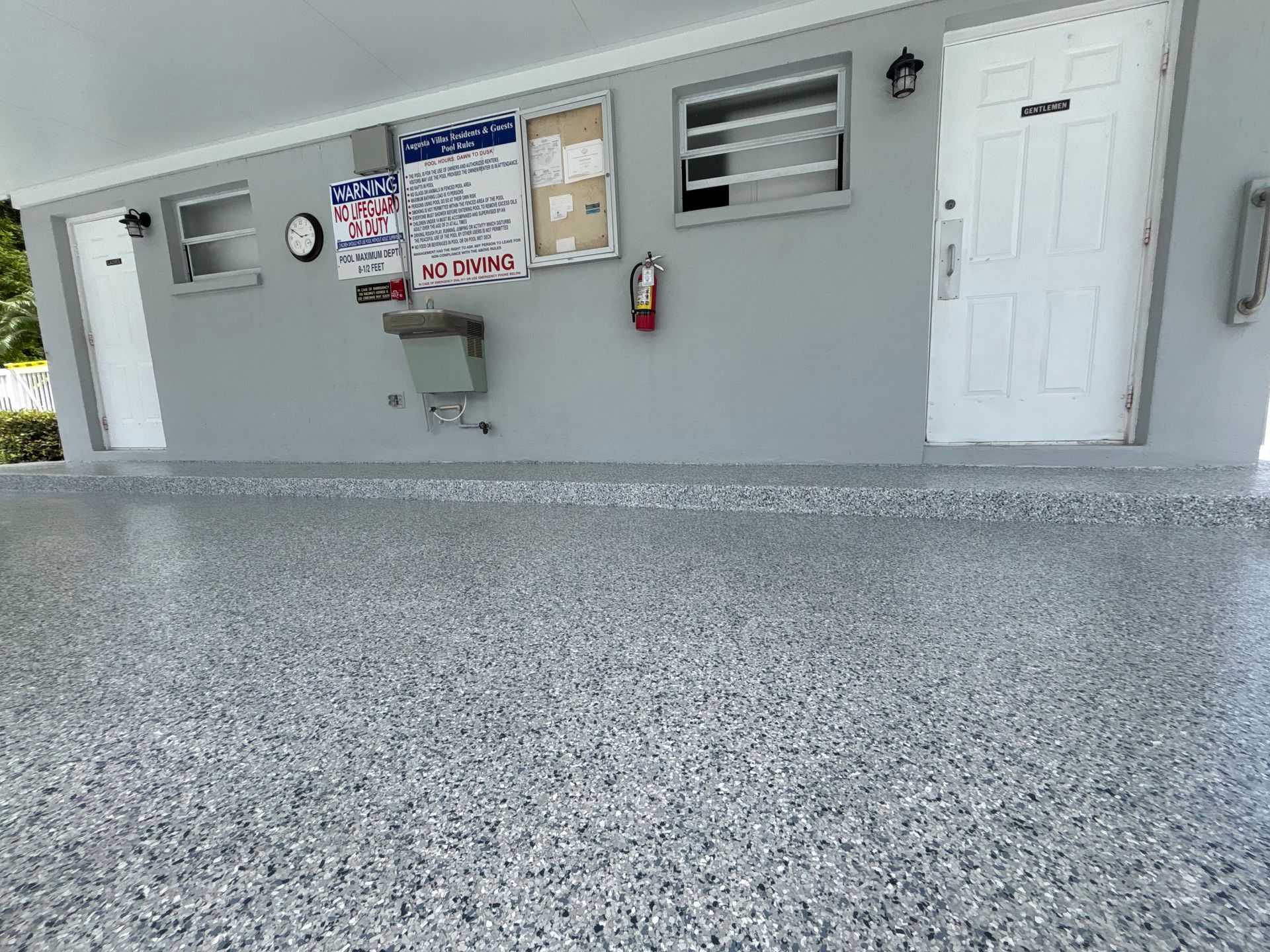 Gray building wall with bulletin boards, wall-mounted sink, fire extinguisher, and white door behind a speckled patio.