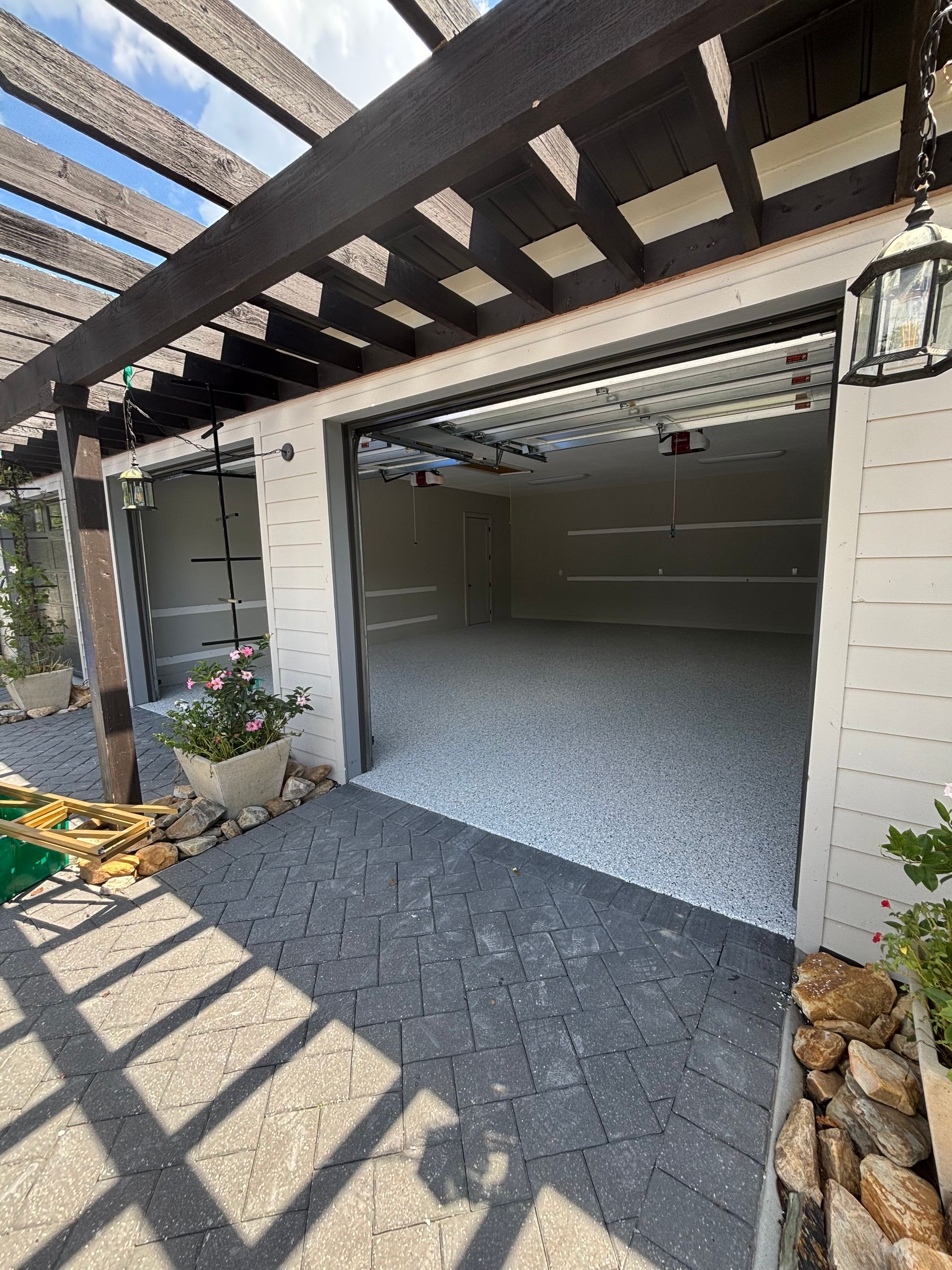 Open garage with gray paver driveway and dark wood pergola casting striped shadows