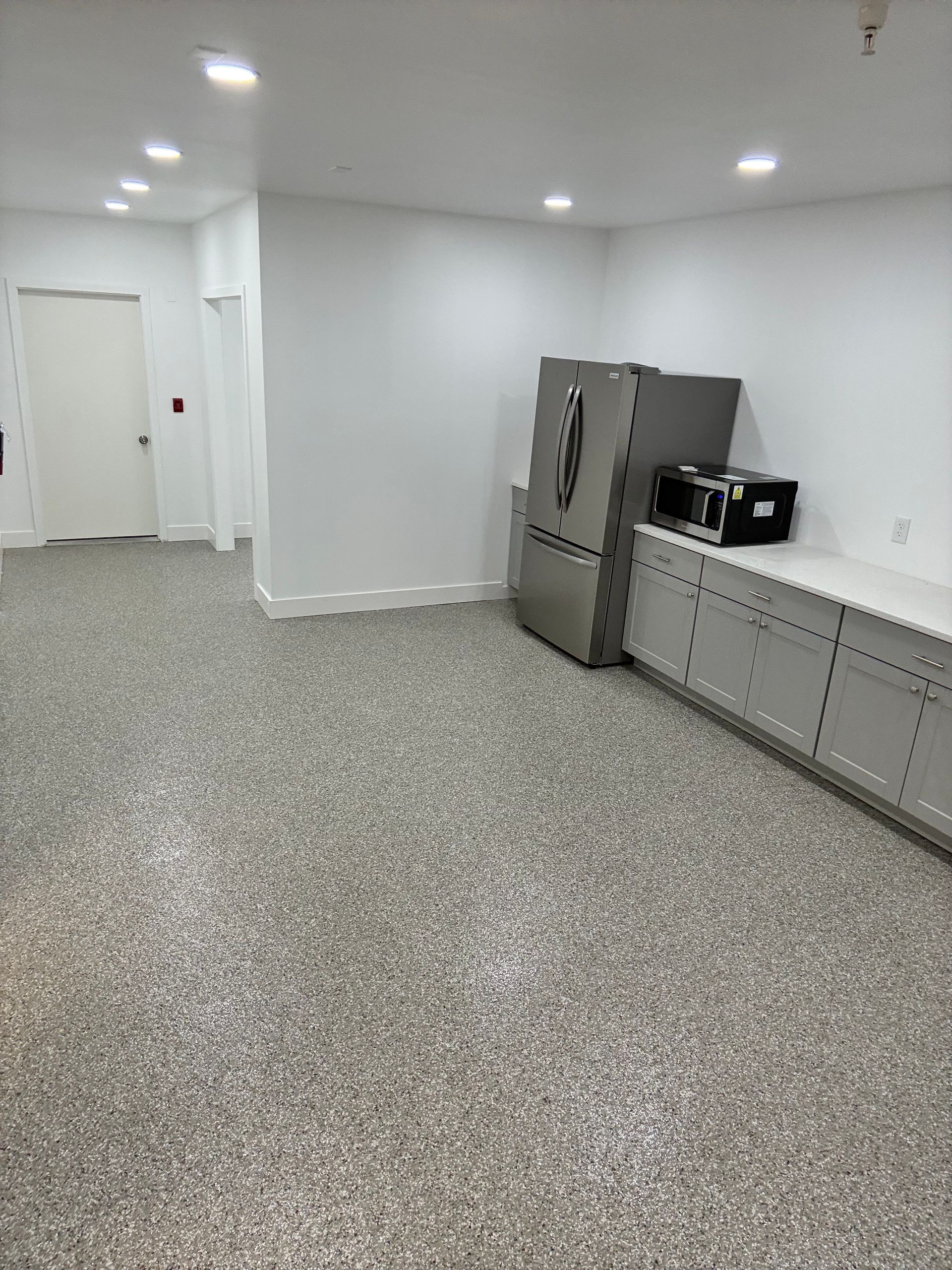 Empty white kitchen with gray speckled floor, stainless fridge, and long countertop cabinets