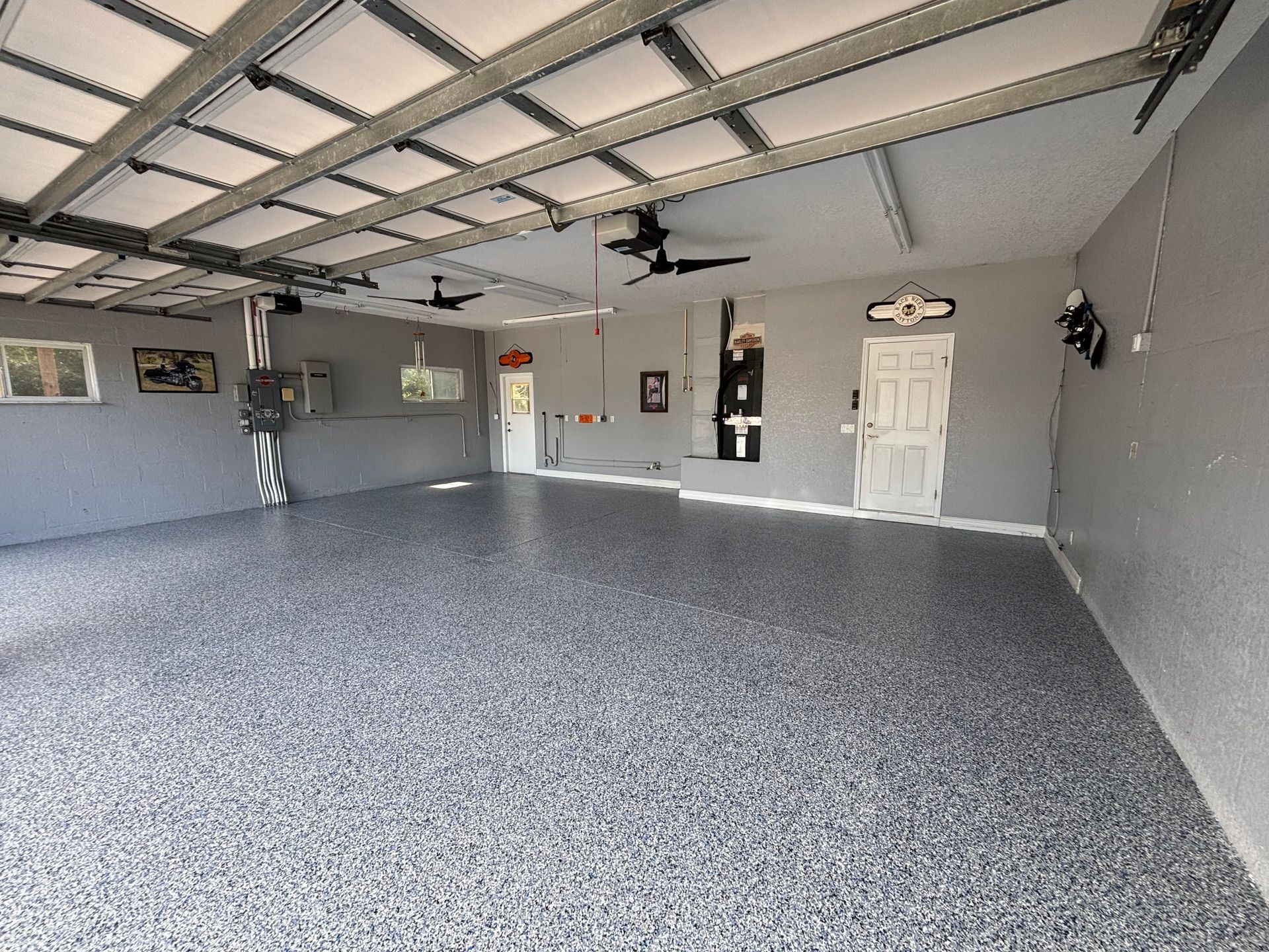 Empty gray garage with speckled floor, white door, wall-mounted tools, and open overhead door with car lift
