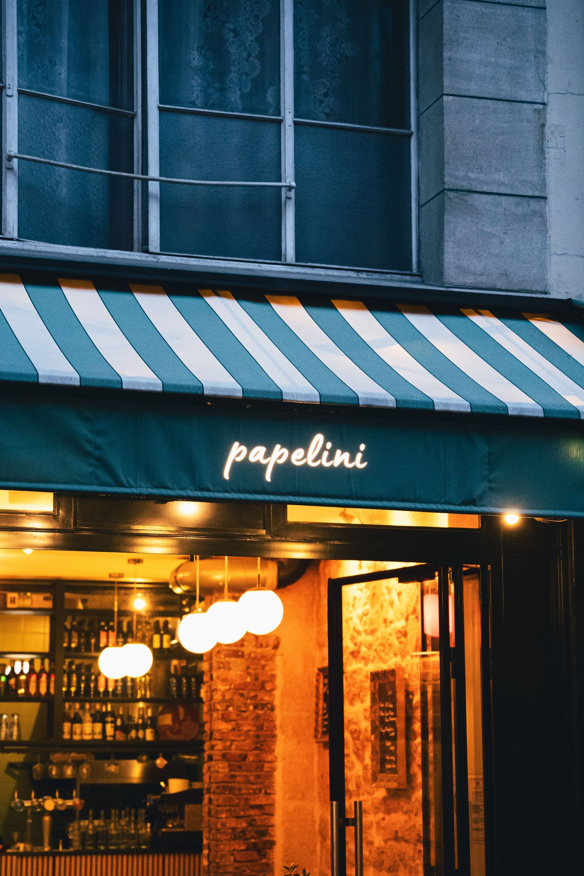 Extérieur du restaurant : enseigne « Papelini » sur un auvent rayé, entrée illuminée et murs en briques.