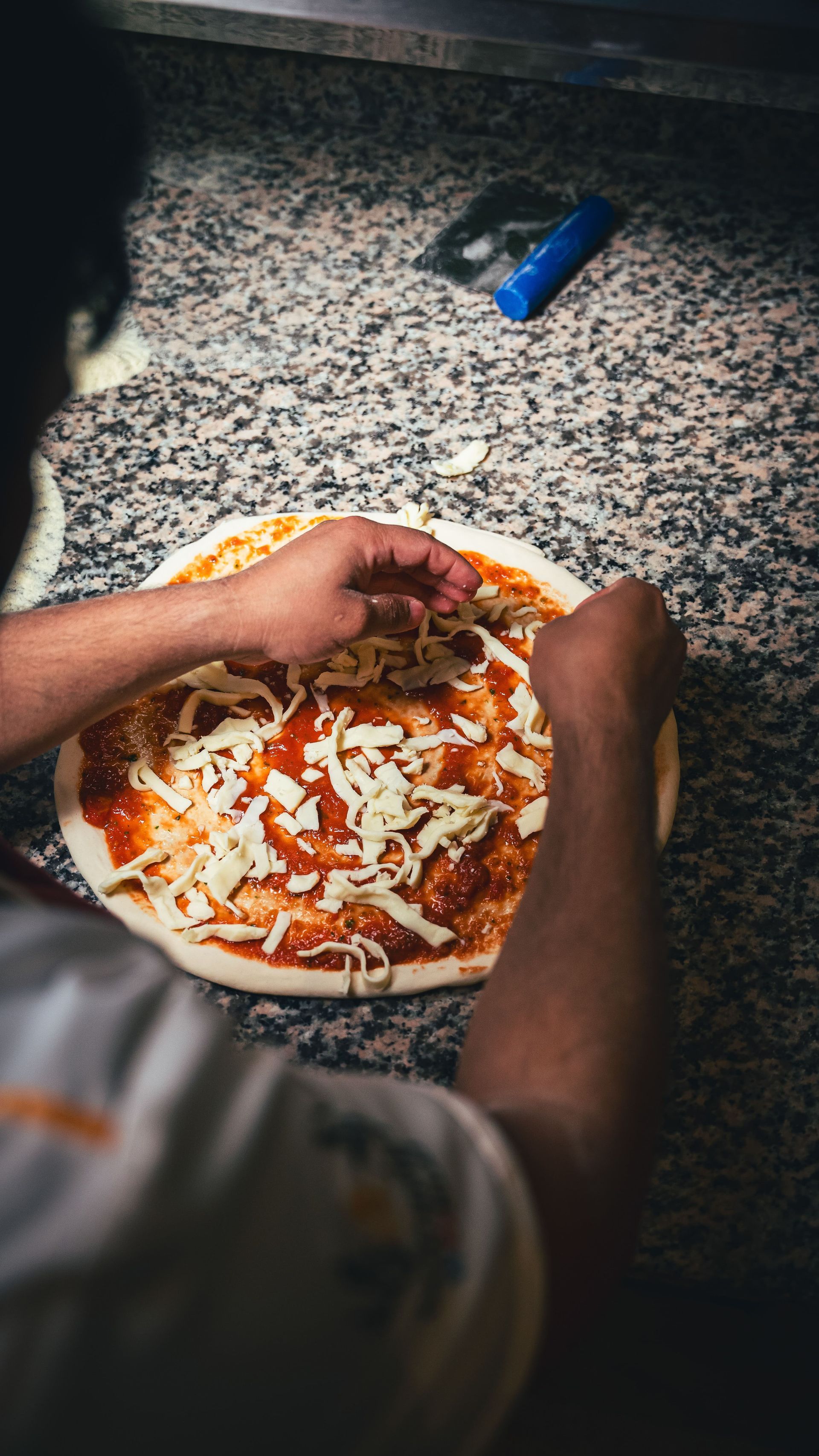 Une personne garnit une pizza de fromage sur un comptoir en granit, avec une roulette à pizza en arrière-plan.