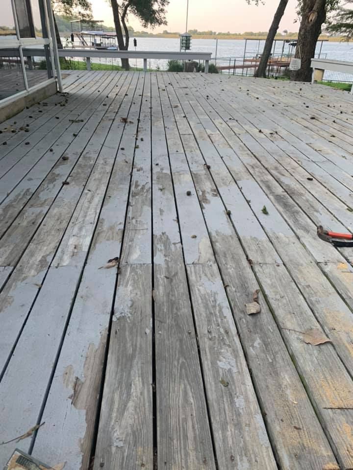 A weathered wooden deck with peeling grey paint, viewed from above, overlooking a body of water with trees in the distance.